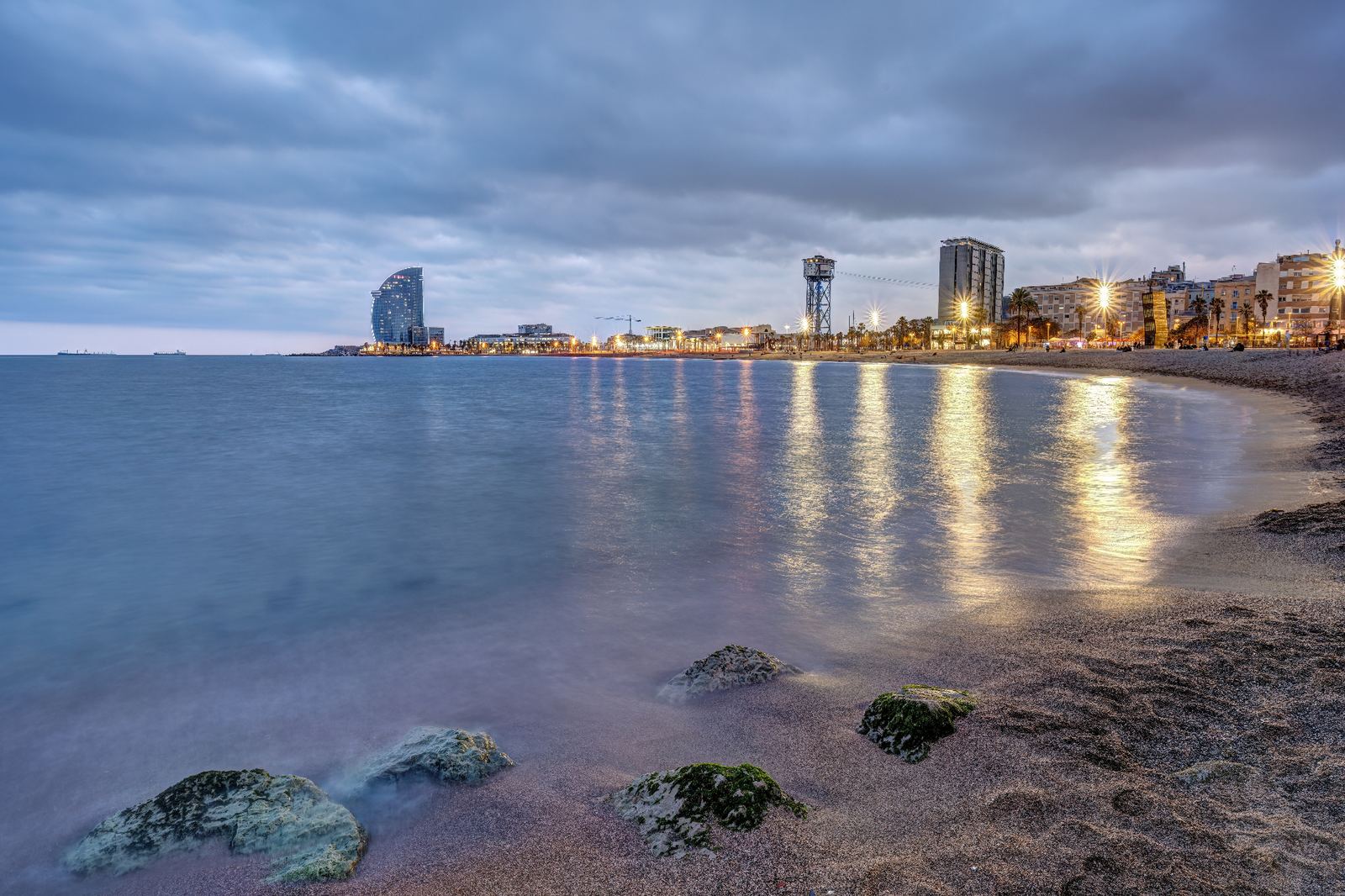 Barceloneta beach at dusk with calm Mediterranean waters, the W Hotel and beachfront lights reflecting on the shoreline