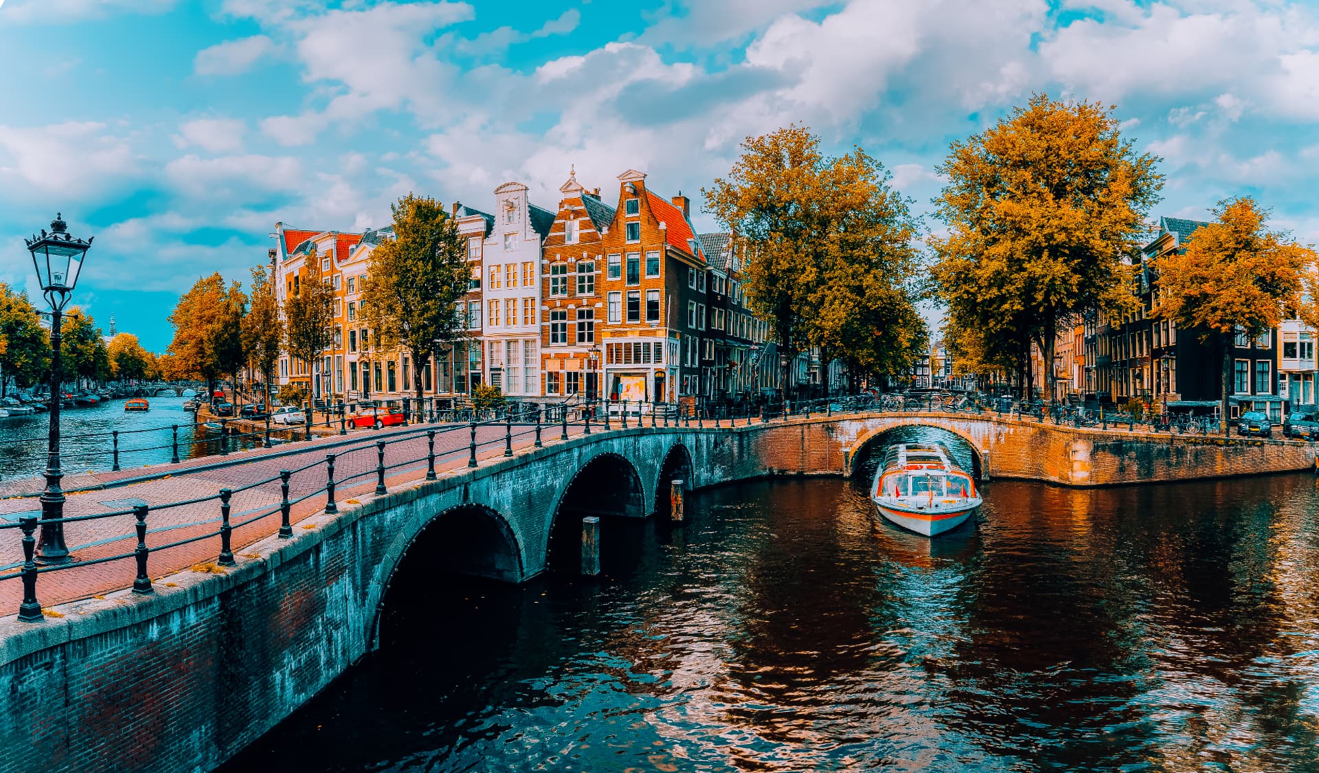 A tour boat passing under a brick bridge on Amsterdam's canal ring with gabled canal houses along the quay