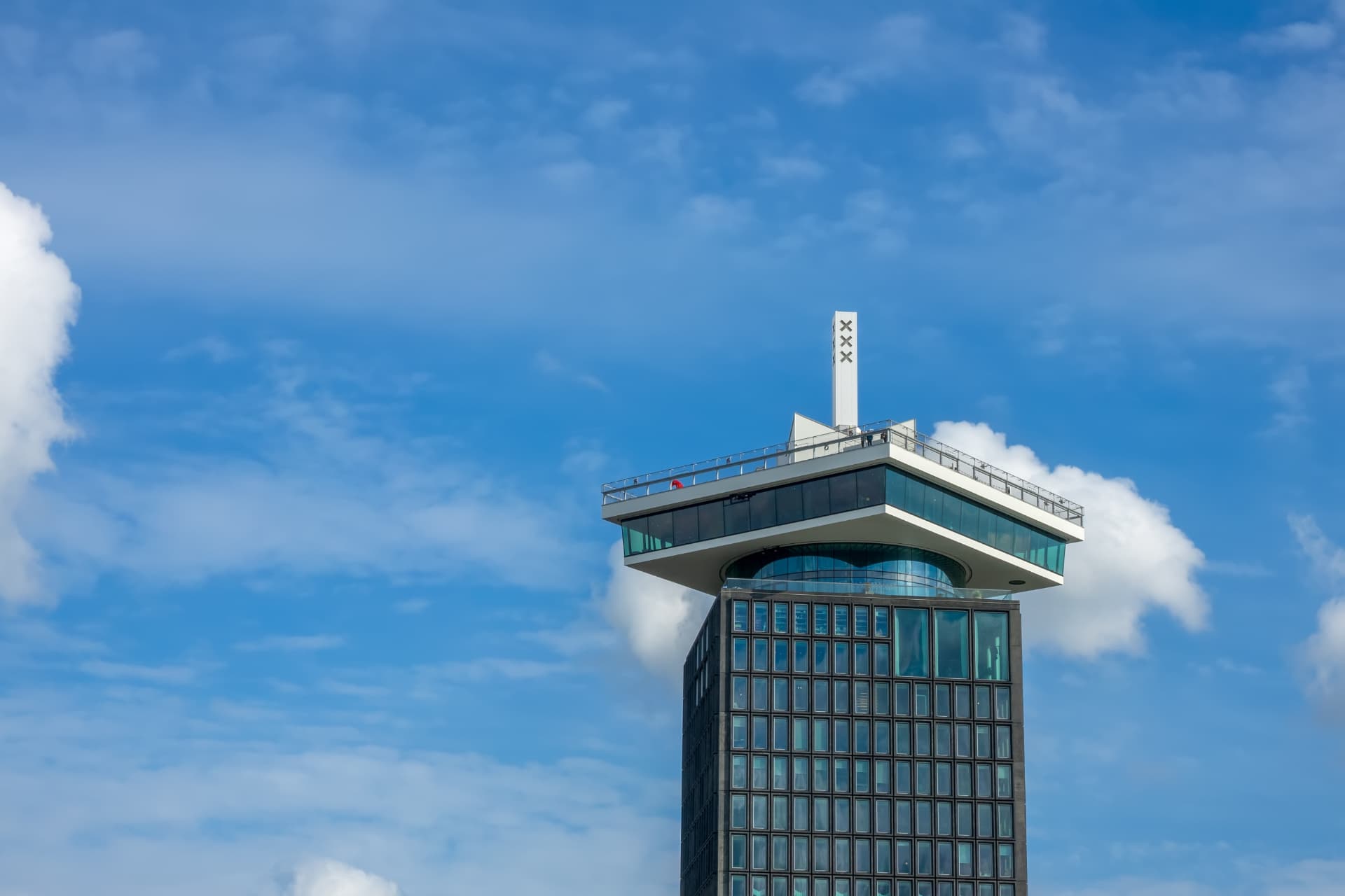 The crown-shaped observation deck of the A'DAM Lookout against a blue sky, with the white mast carrying Amsterdam's three crosses