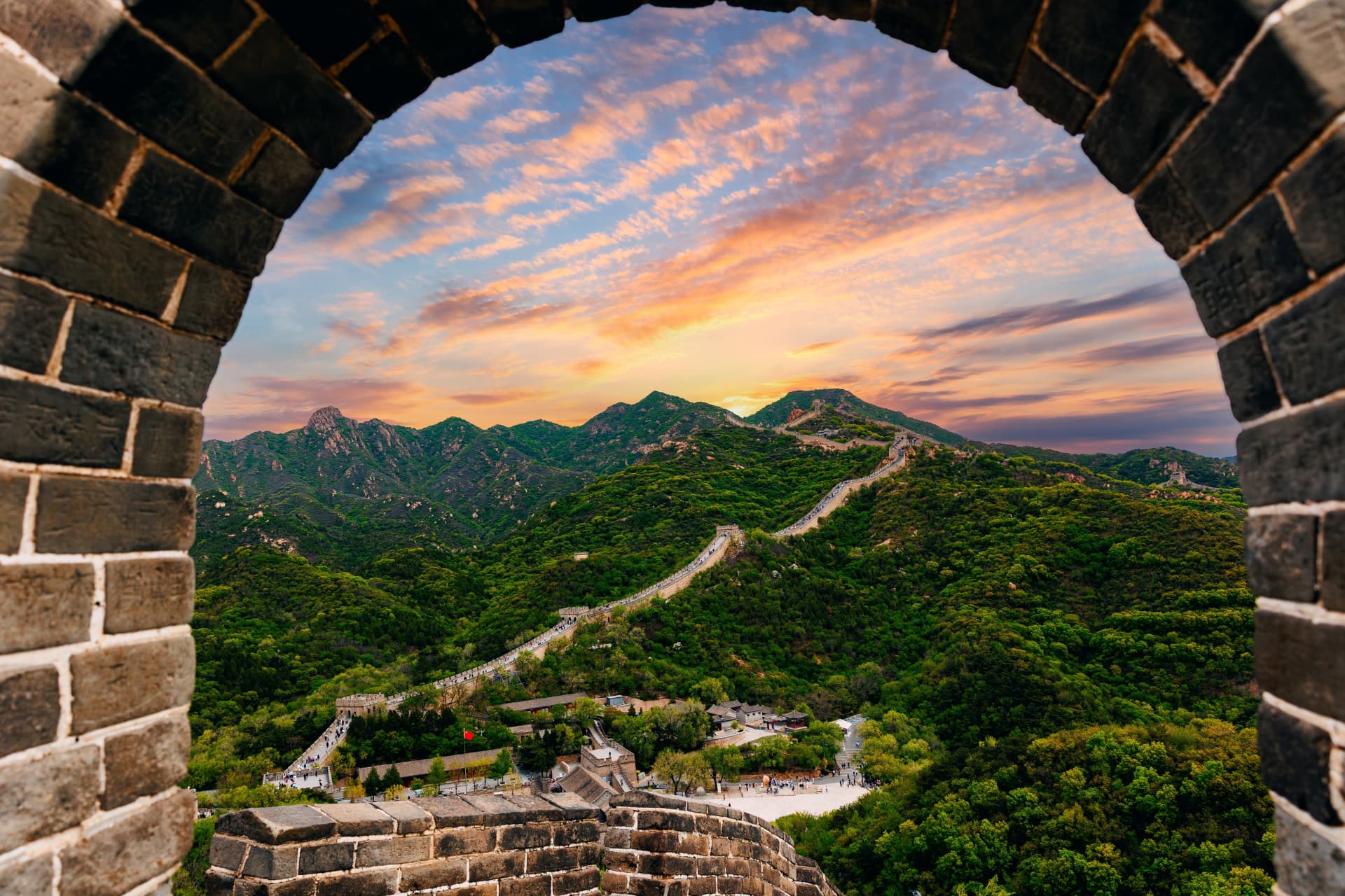 The Great Wall of China snaking across green mountain ridges at sunset, framed by a brick watchtower window
