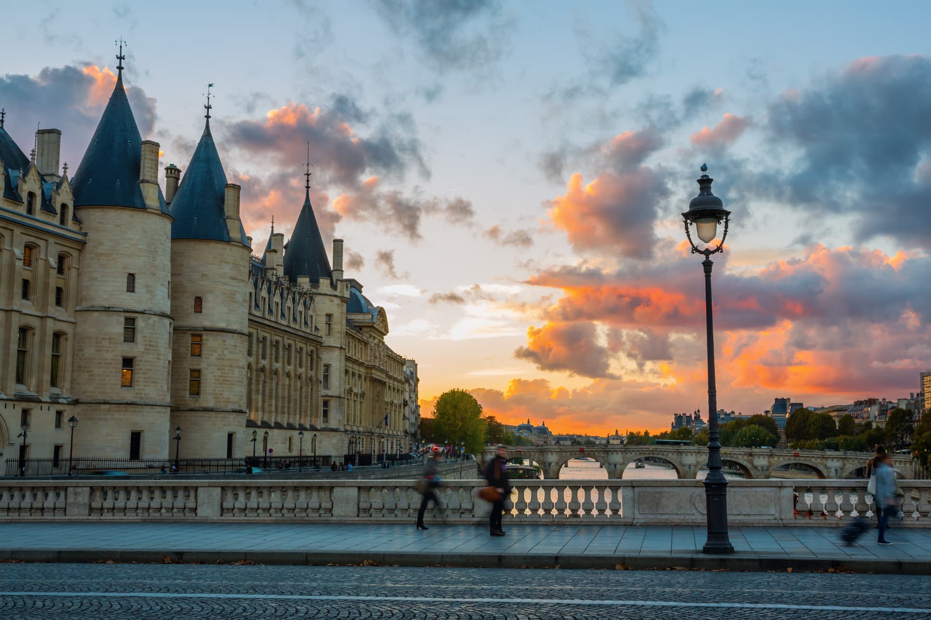 The conical towers of the Conciergerie above the quay with pedestrians and a street lamp at sunset