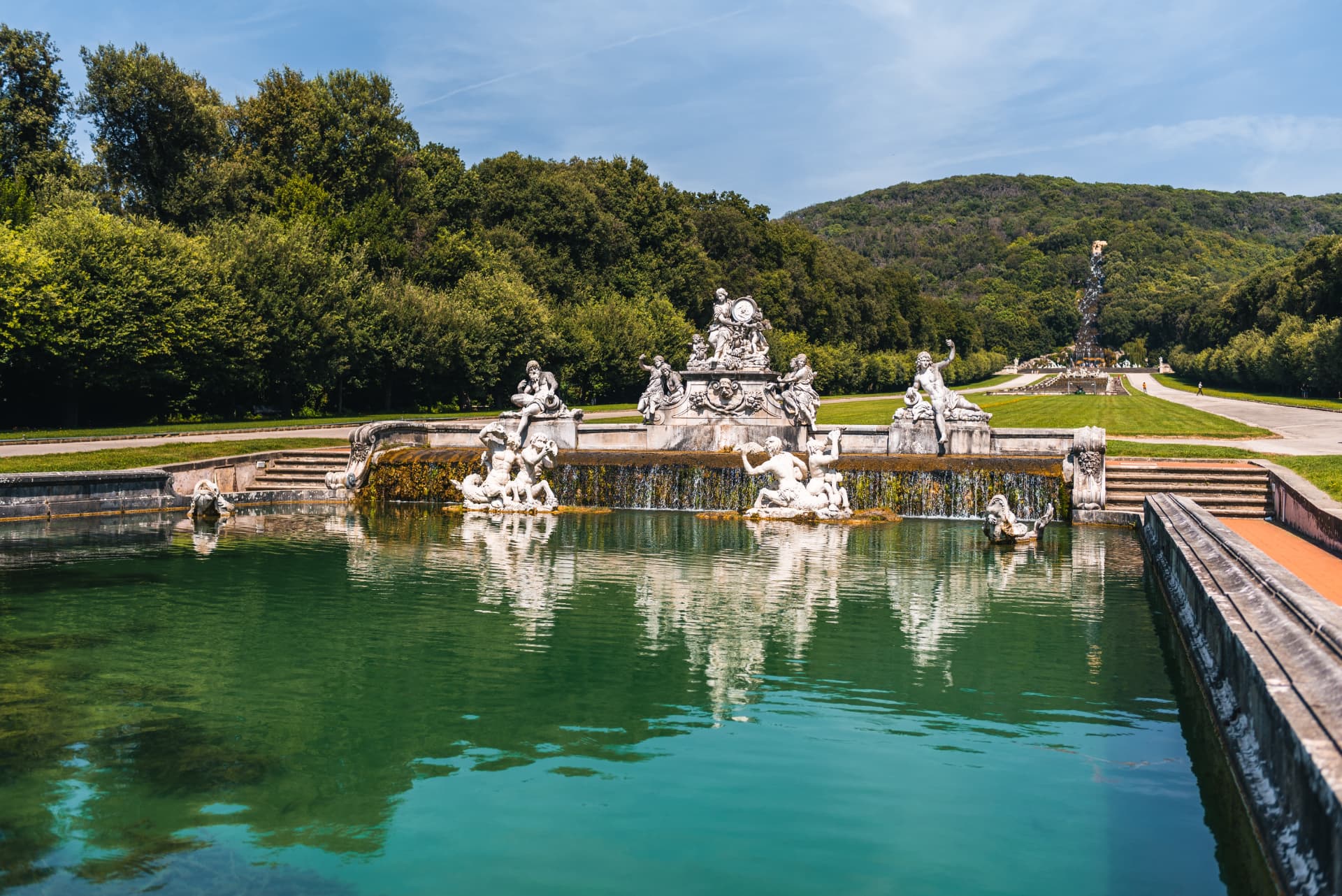 The Fountain of Ceres in the Royal Park of Caserta with white statues and a green pool, the waterfall visible on the hill behind