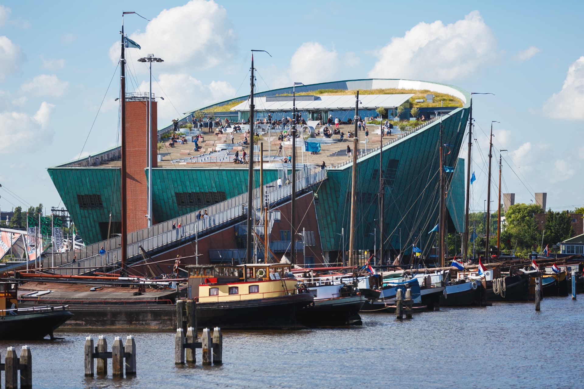 Visitors relaxing on the sloping rooftop terrace of the NEMO Science Museum in Amsterdam with historic sailing barges in the foreground