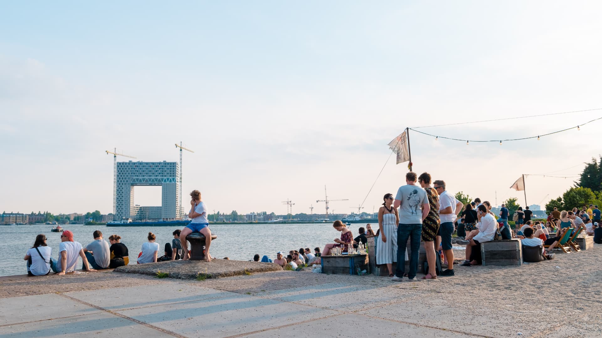 Visitors sitting by the water at NDSM Wharf in Amsterdam with boats passing on the IJ