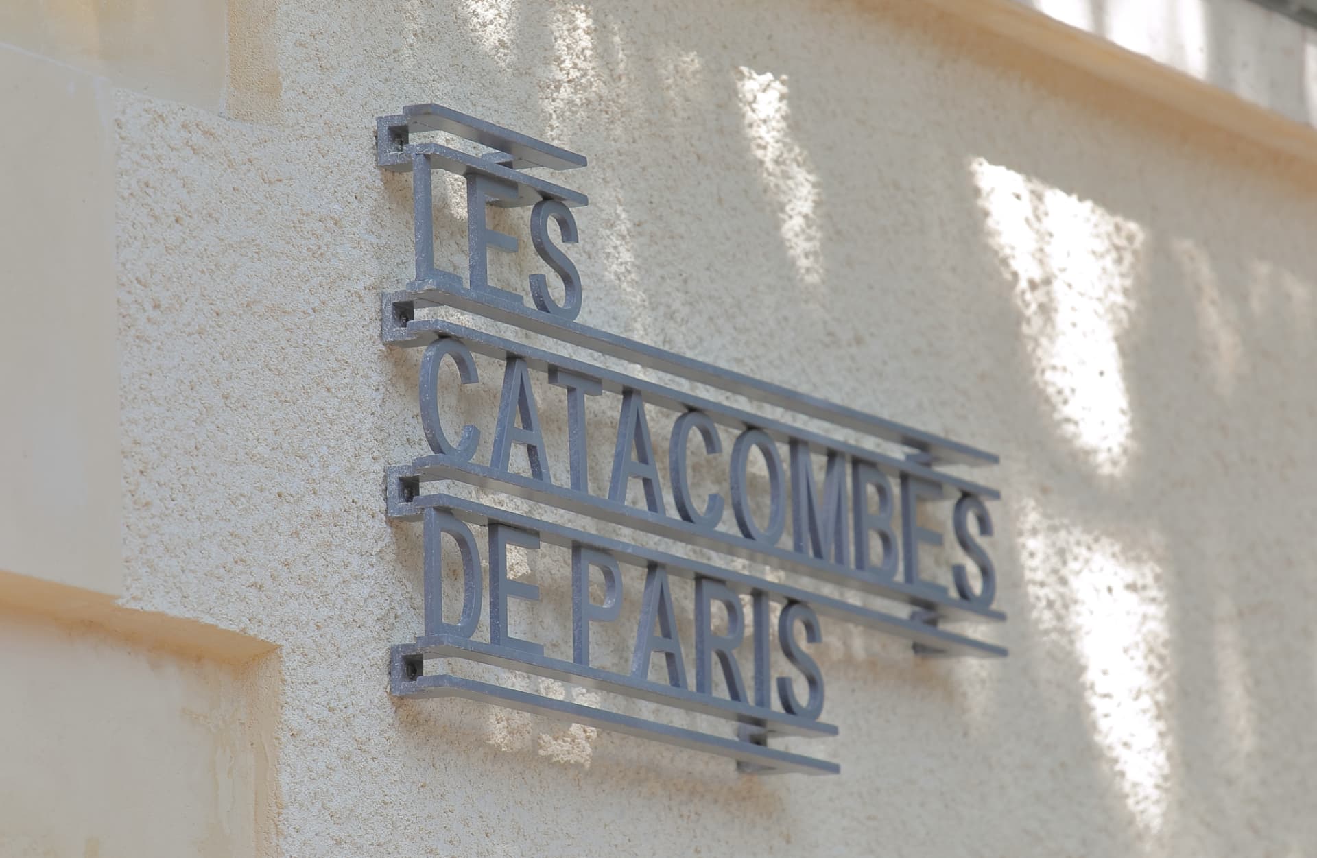 The metal 'Les Catacombes de Paris' sign on the stone wall of the entrance building at Denfert-Rochereau