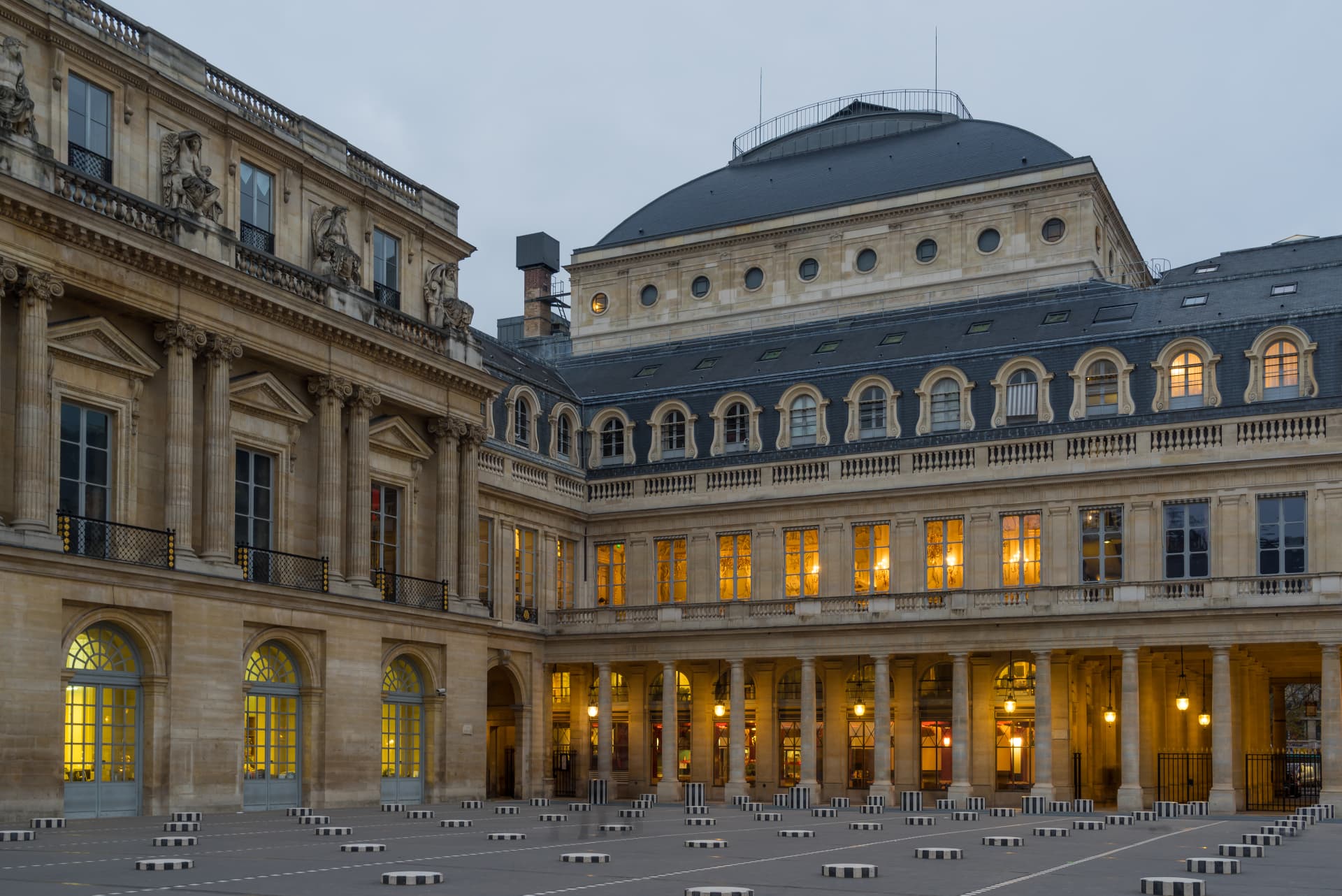 Palais Royal courtyard at dusk with warm light in the arcade windows and Buren columns in the foreground