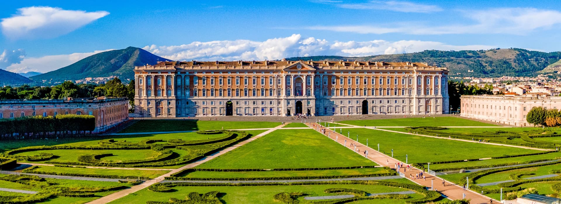 Front of the Royal Palace of Caserta with its long orange and stone facade behind green lawns and patterned hedges