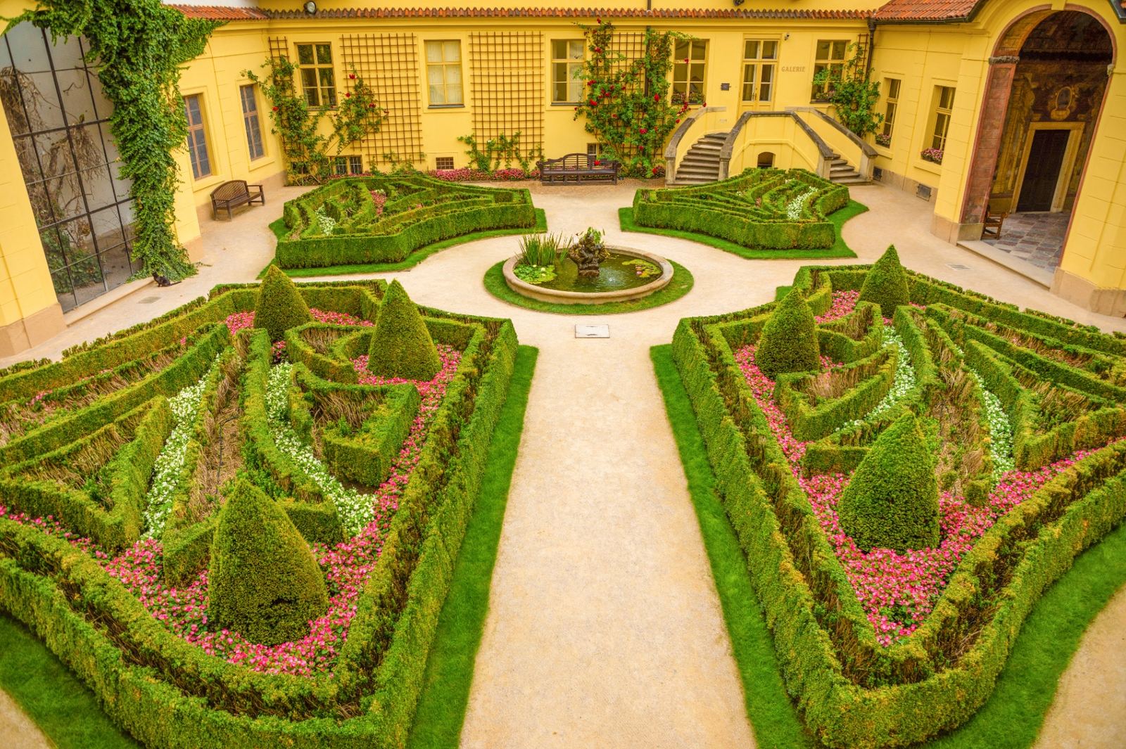 Vrtba Garden in Prague: a baroque courtyard garden with geometric boxwood hedges, pink flower beds, a central fountain and a yellow historic palace behind