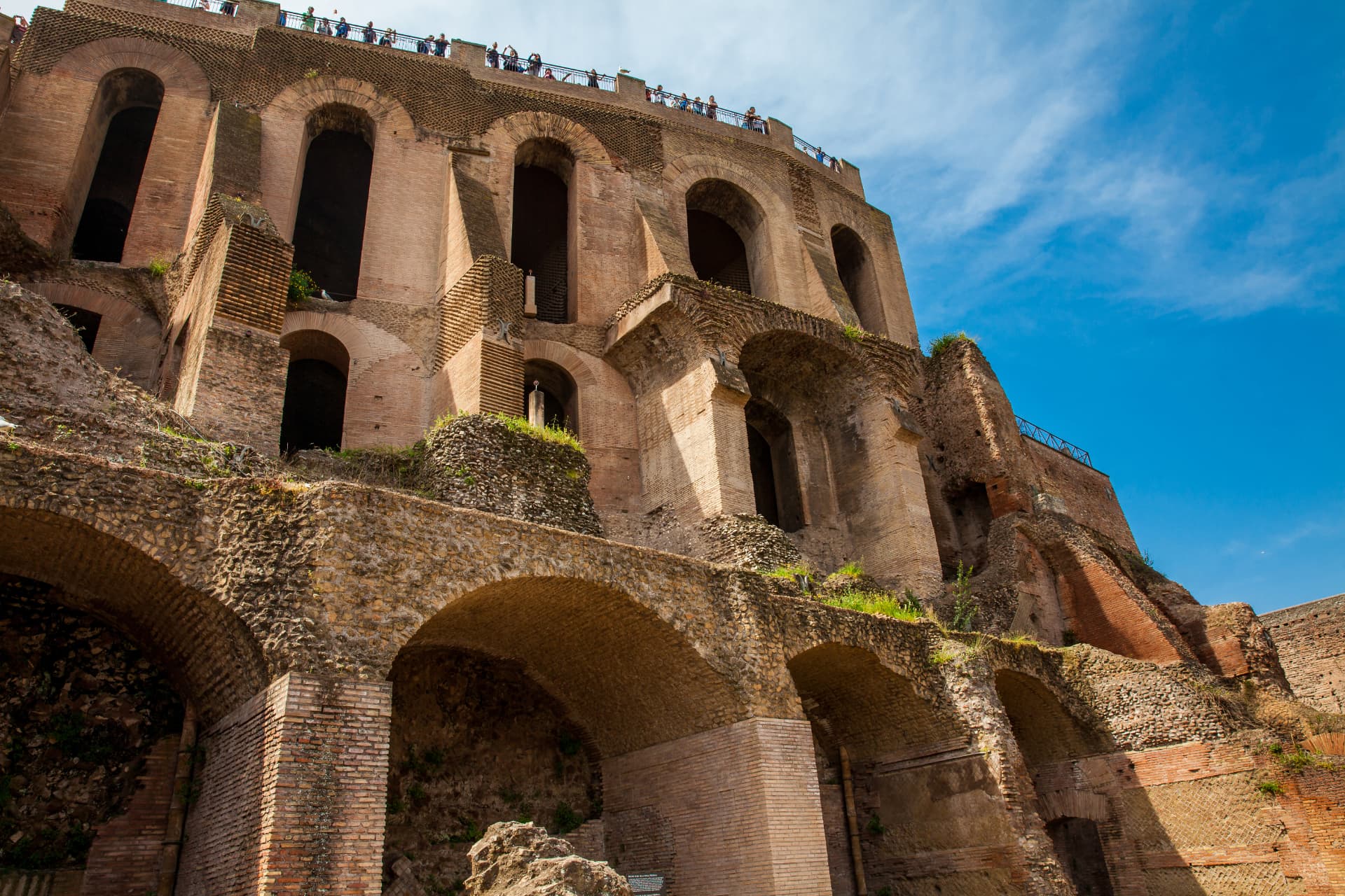 Tall brick arcades of the Domus Tiberiana rising above the Roman Forum with visitors on the terrace