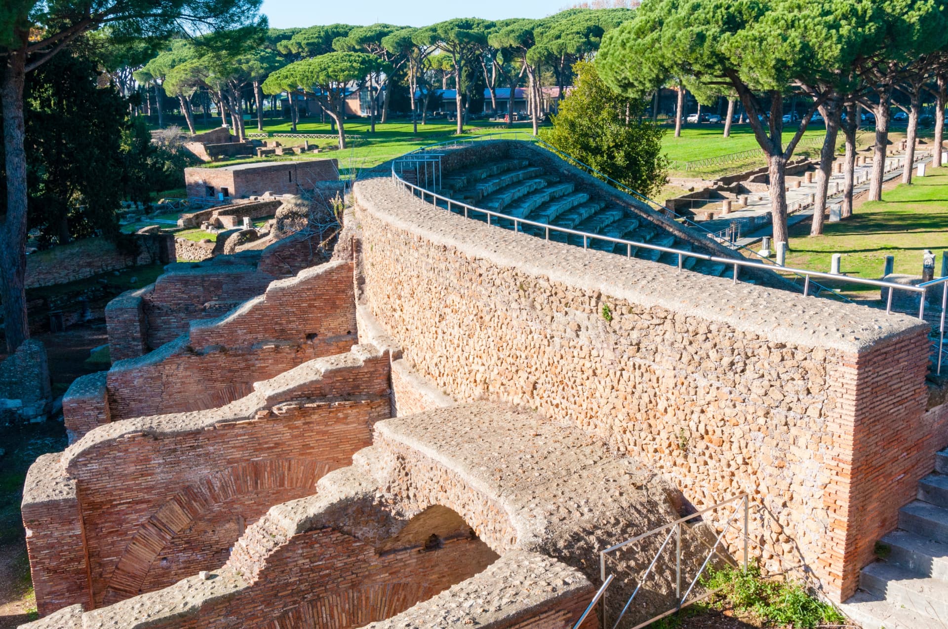 View over the brick ruins of Ostia Antica towards the theatre's curved stone seating, framed by umbrella pines