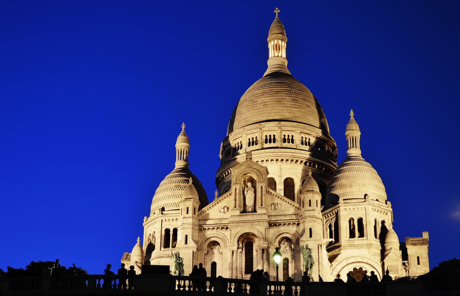Sacré-Cœur Basilica floodlit against a deep blue evening sky, with visitors silhouetted along the balustrade in front