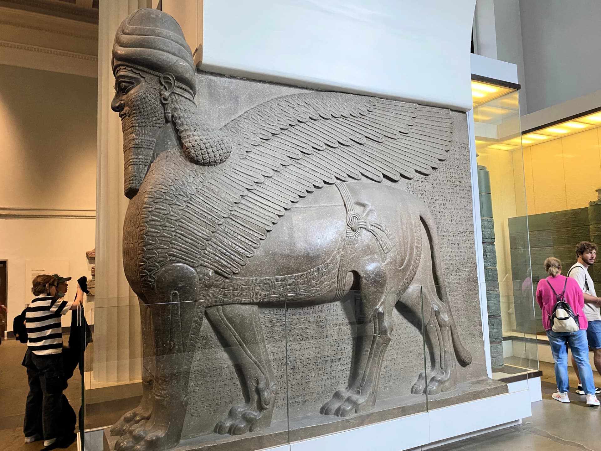 A large Assyrian winged bull relief inside the British Museum with visitors nearby