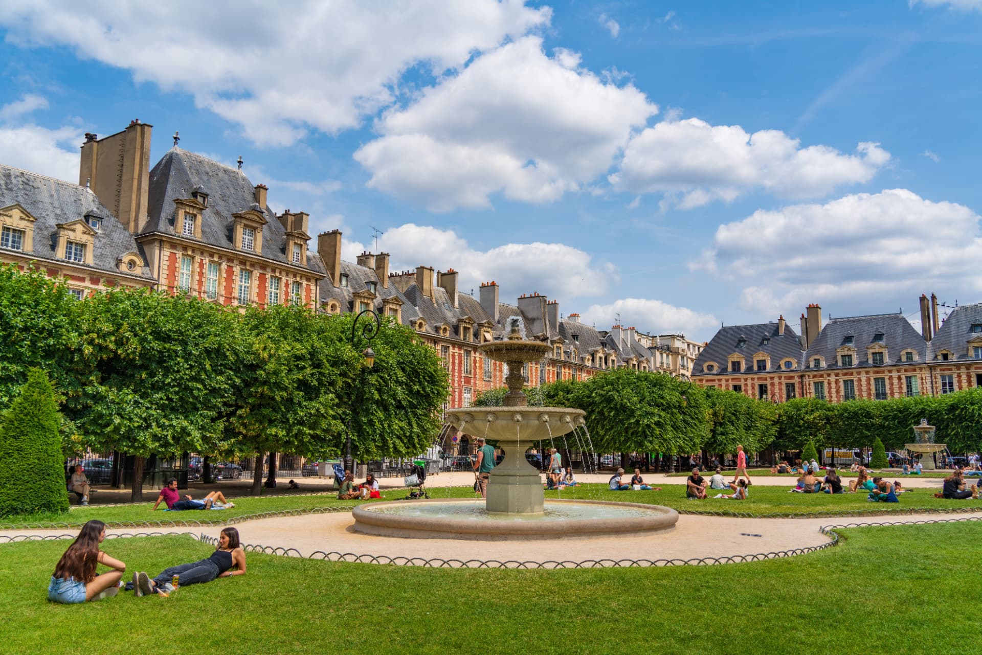 People relaxing on the lawns of the Place des Vosges garden around the central fountain on a summer day