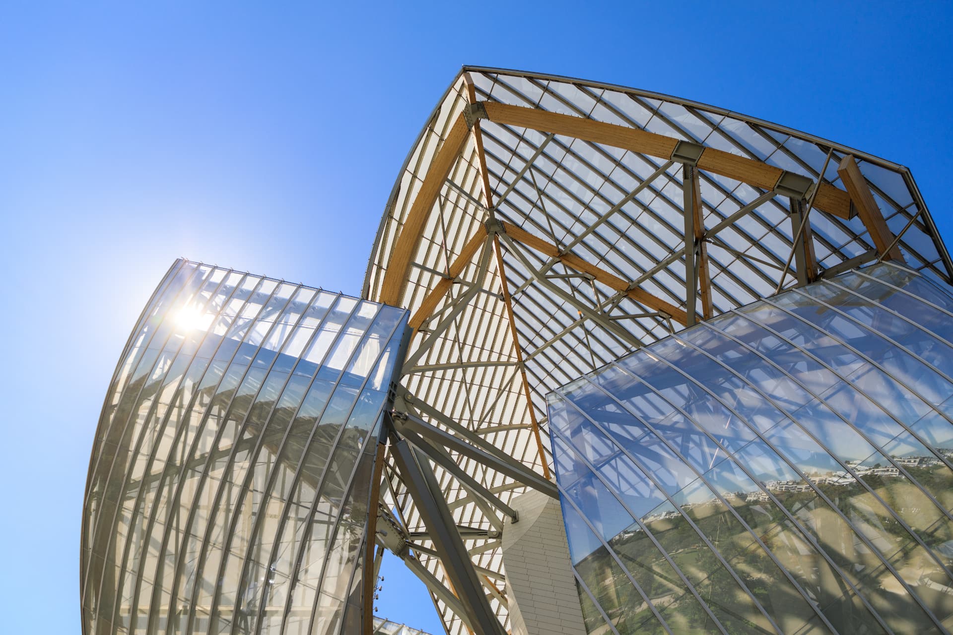 Looking up at the curved glass sails of the Fondation Louis Vuitton against a deep blue sky, with their wooden and steel supporting beams visible