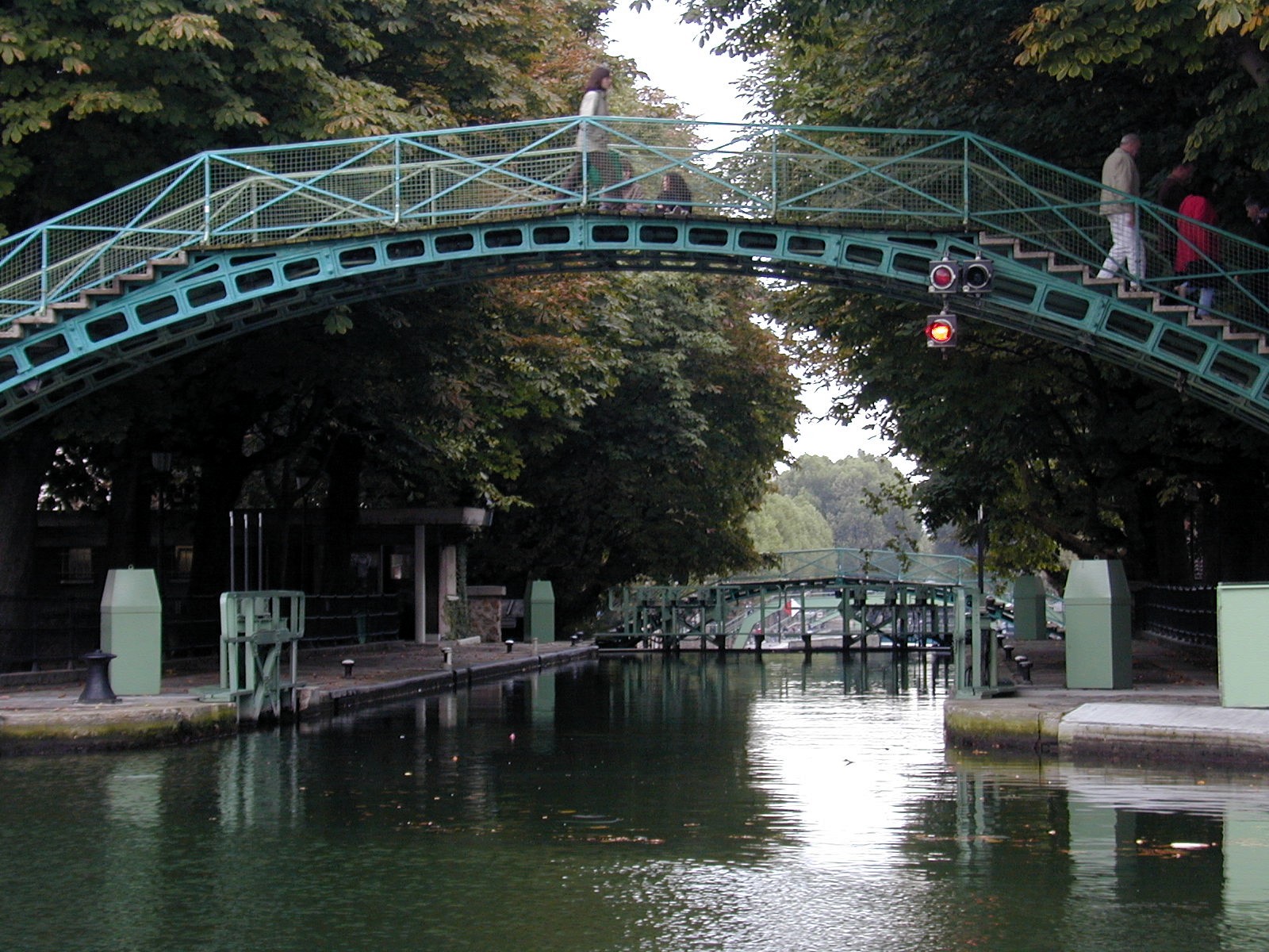 A green iron footbridge arching over the Canal Saint-Martin in Paris, with a lock and a second bridge visible beyond