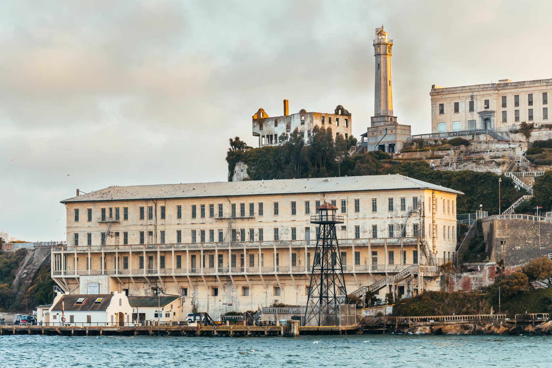Alcatraz Island seen from the water at dusk, showing the barracks building, guard tower, lighthouse and the ruined warden's house above