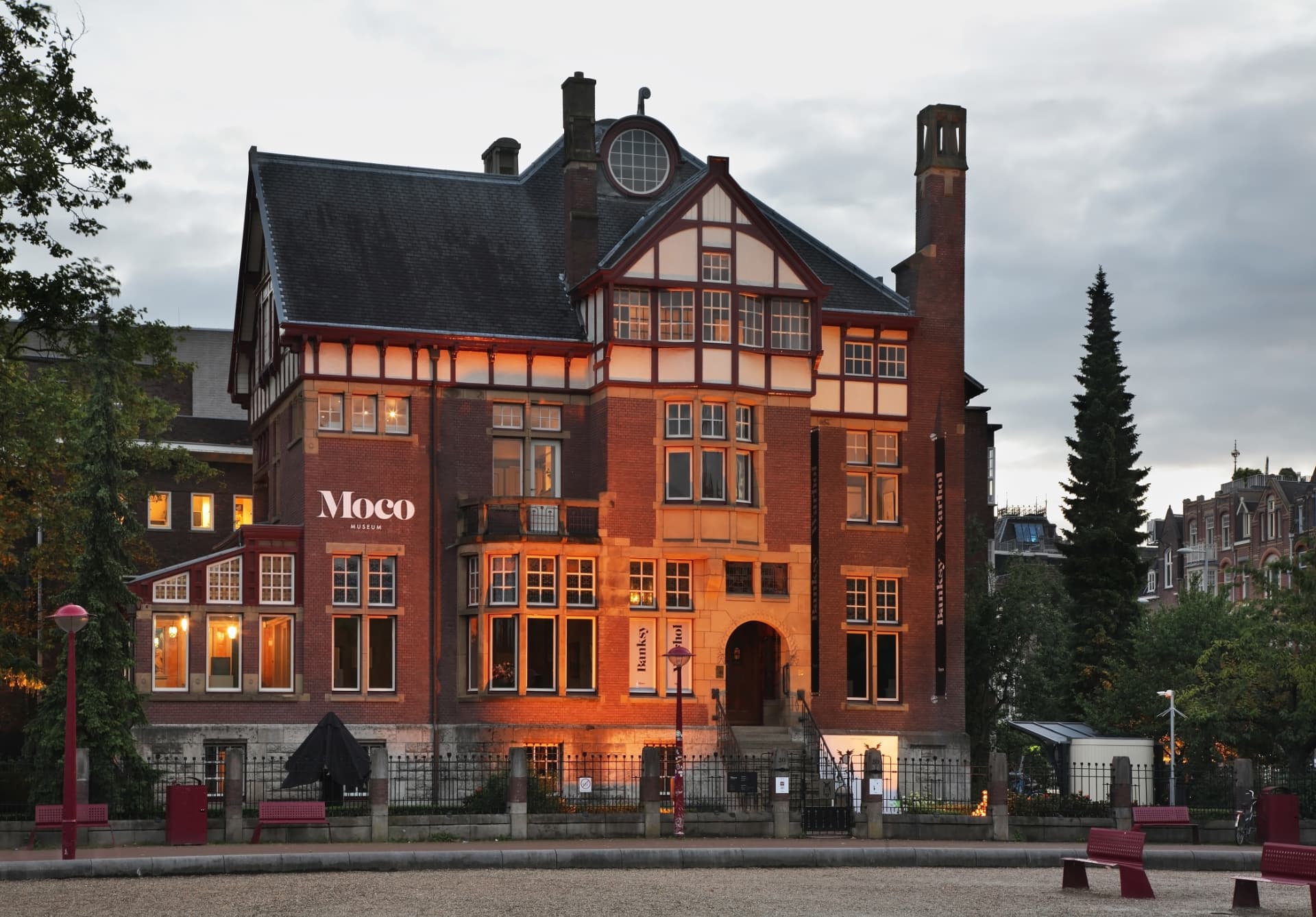 The Moco Museum villa glowing warmly at dusk, with its red-brick facade and timber gables lit from inside