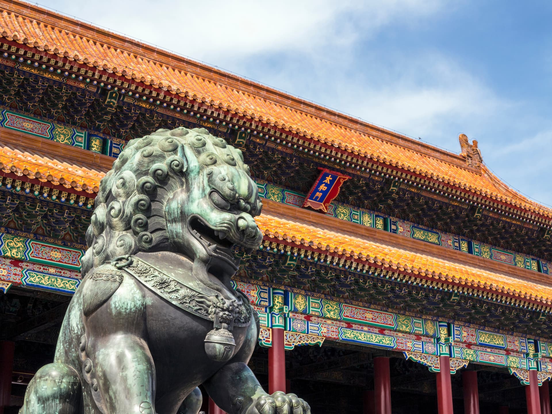 Bronze guardian lion in front of ornate painted beams and golden Forbidden City roof tiles