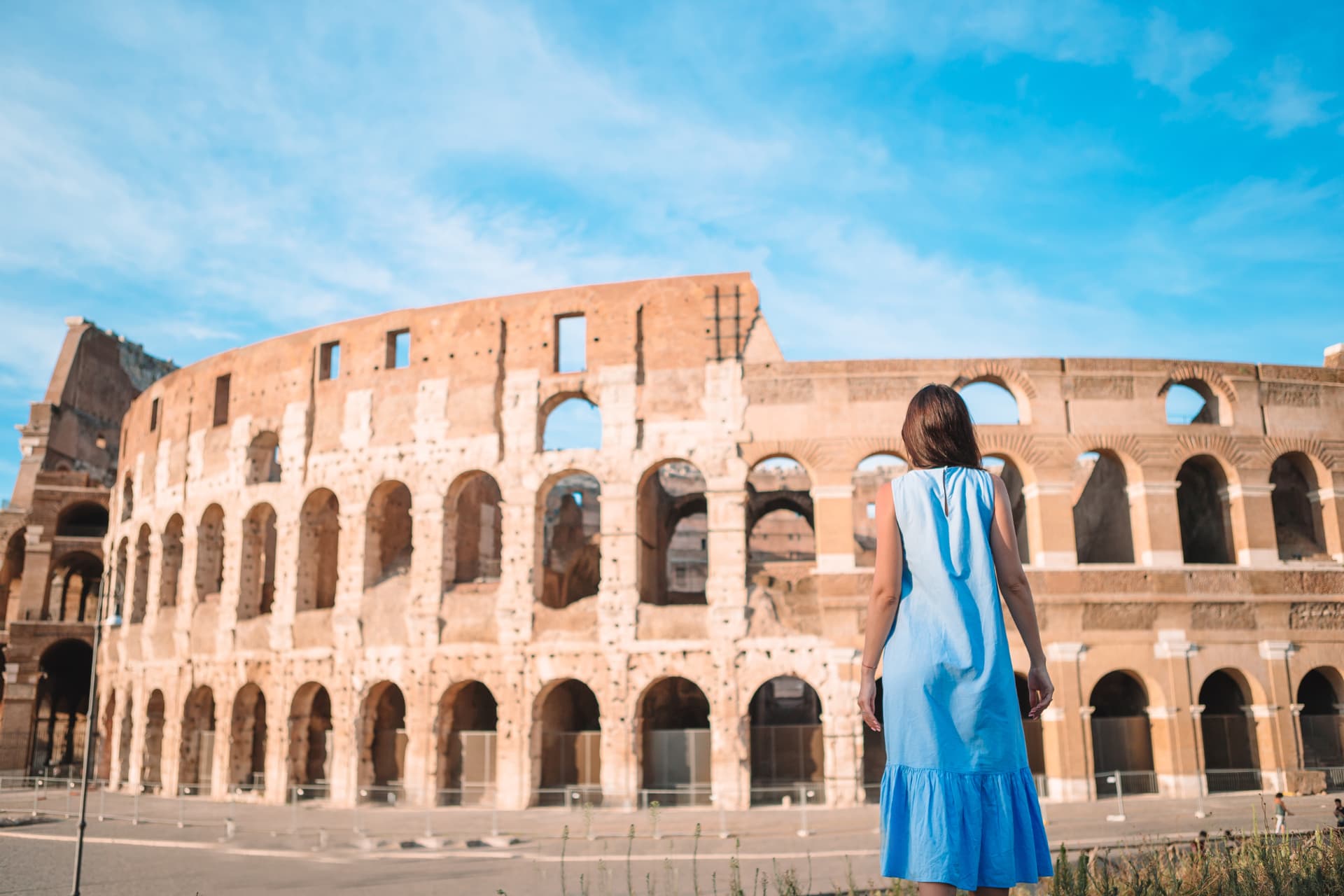 Visitor in a blue dress standing in front of the Colosseum exterior walls in Rome on a clear morning
