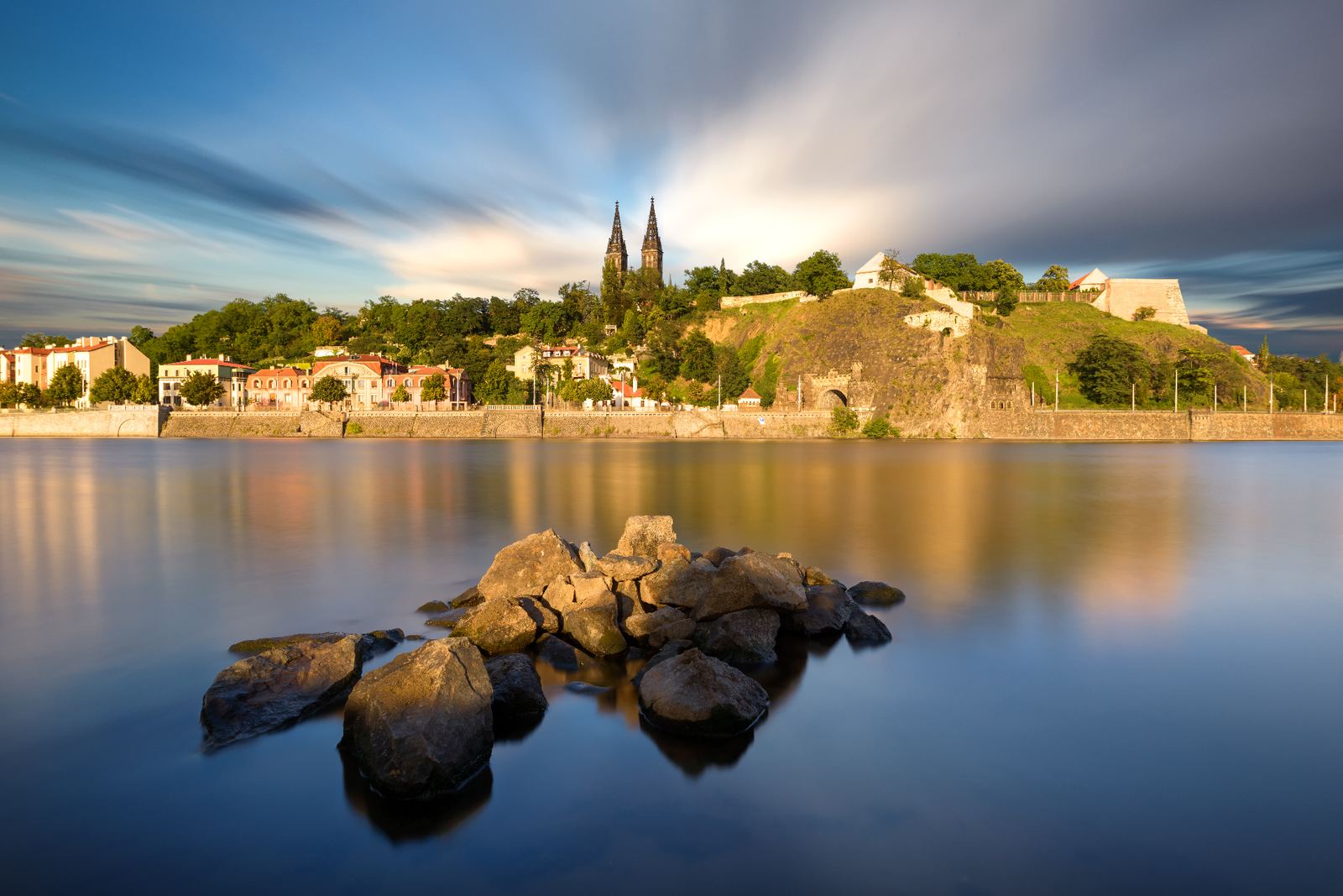 Vyšehrad fortress and the twin neo-Gothic spires of the Basilica of St Peter and St Paul rising above the Vltava River at golden hour, with rocks reflected in the still water