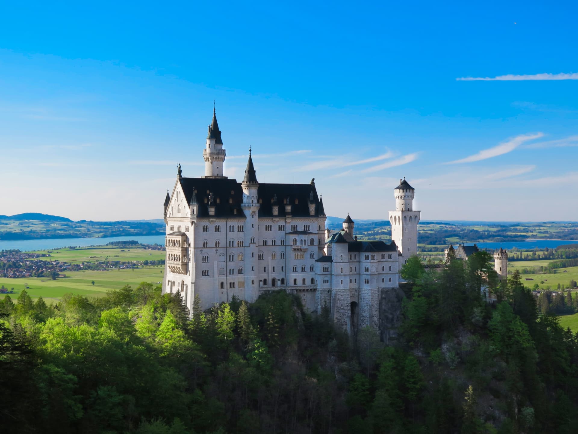 Neuschwanstein Castle on its wooded crag above the Bavarian plain, with the Forggensee lake and green fields behind it