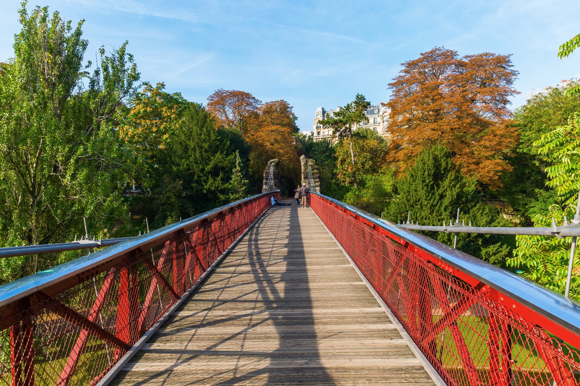 The red iron suspension bridge of the Parc des Buttes-Chaumont seen from its wooden deck, with autumn trees and Haussmann buildings beyond
