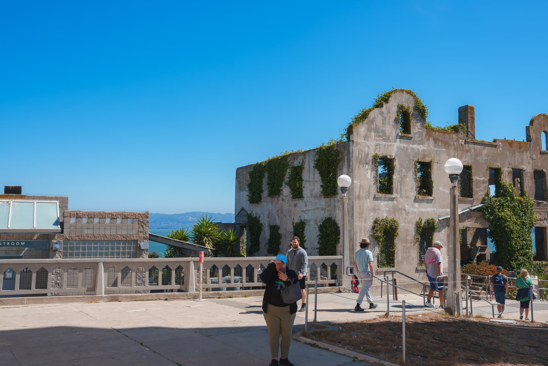 The ivy-covered ruined shell of a building on Alcatraz Island with visitors walking past on a bright day