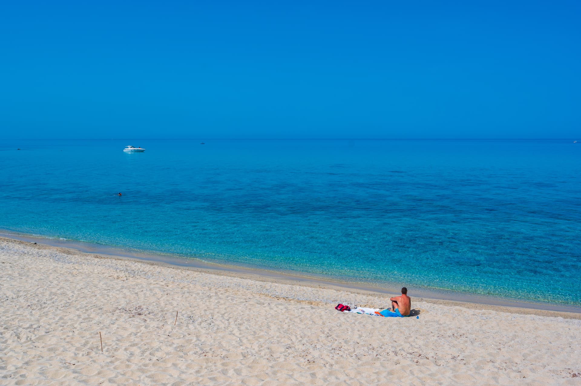 A wide sandy beach with clear blue water and a single swimmer, a boat on the horizon