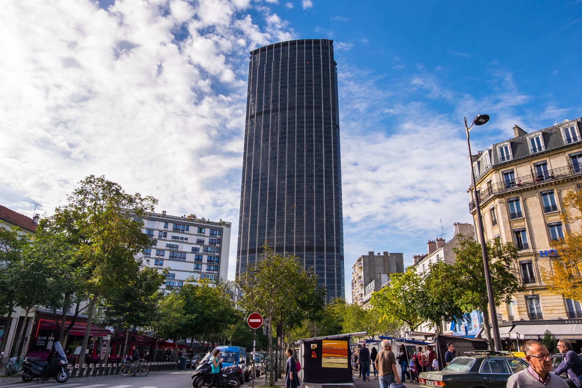 The Montparnasse Tower seen from street level with cafés, market stalls and Haussmann buildings in the foreground