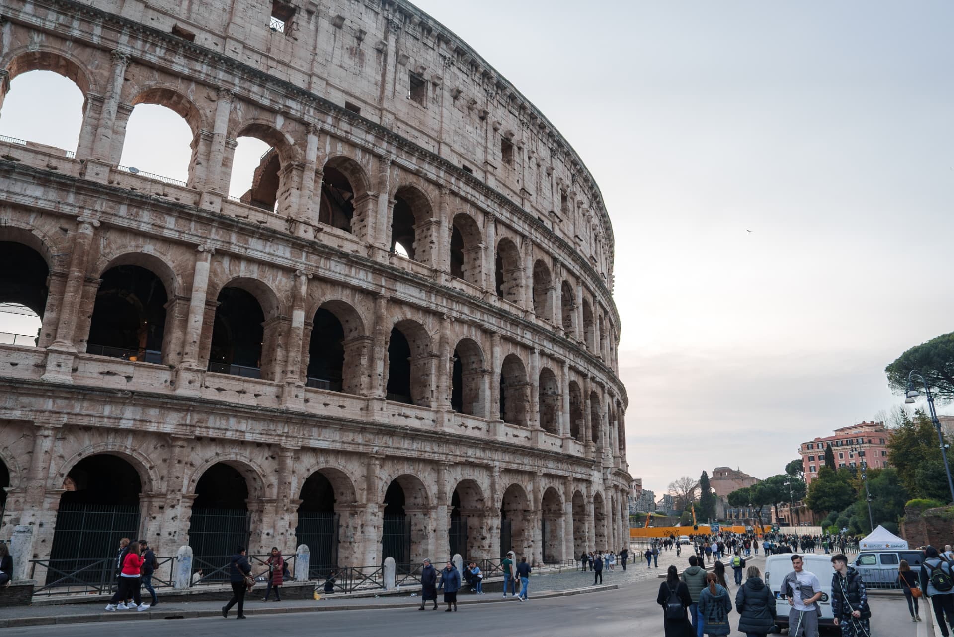 Street view of the Colosseum exterior with visitors walking along Via Sacra in Rome