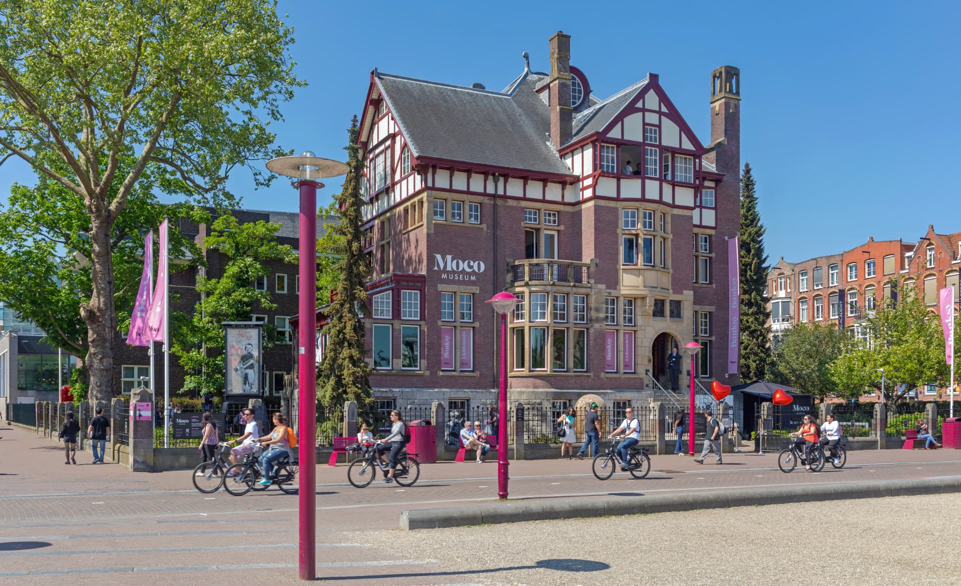 Cyclists passing the Moco Museum's pink-flagged villa on the edge of Museumplein