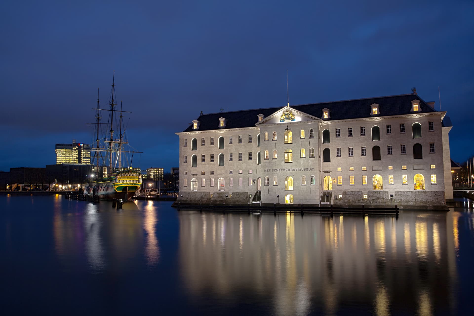 The National Maritime Museum in Amsterdam illuminated at night with the replica tall ship alongside, reflected in the harbour