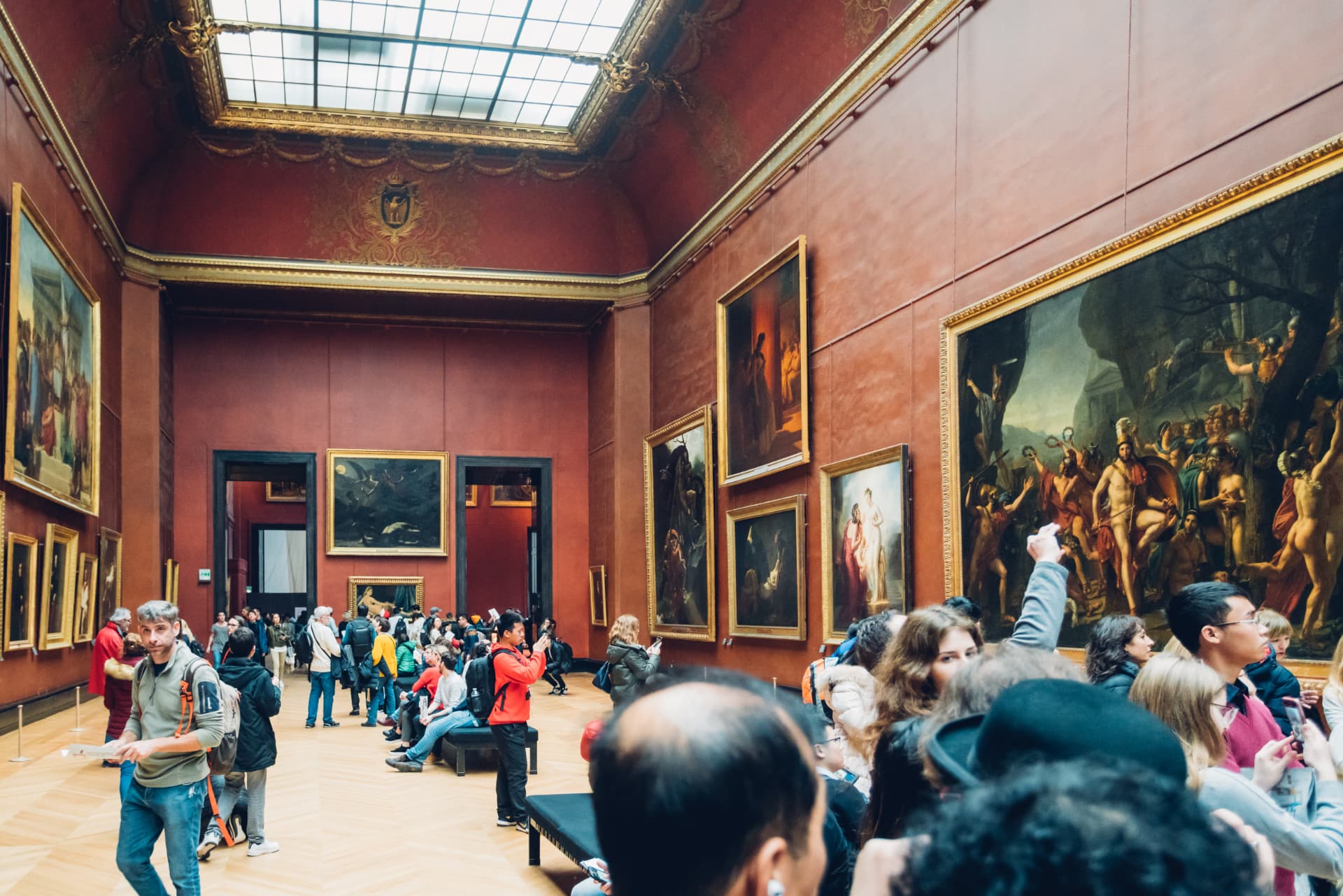 Visitors looking at paintings in a red-walled Louvre gallery