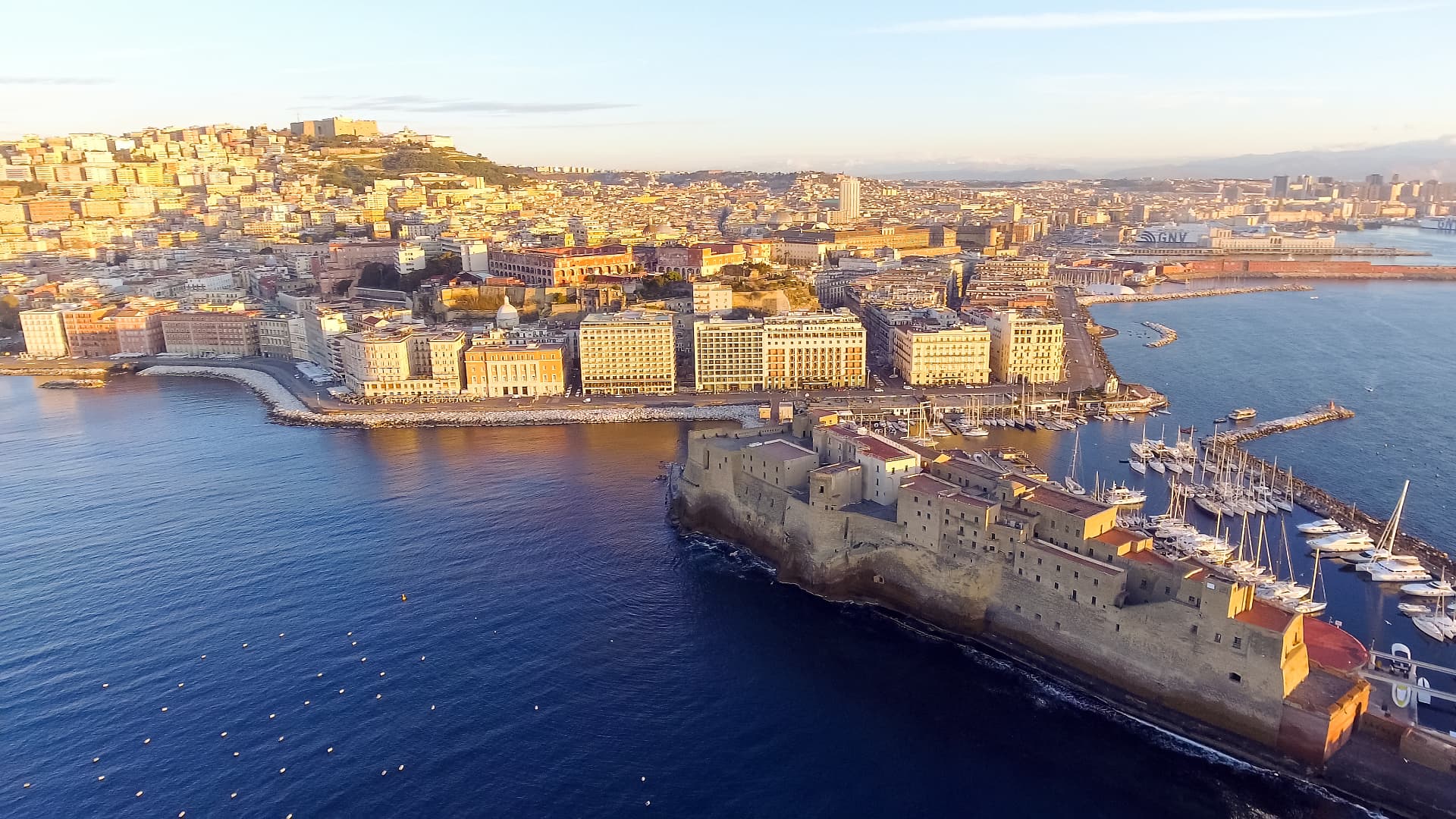 Aerial view of Naples' waterfront at sunset with Castel dell'Ovo on its jetty and the city climbing the hills behind