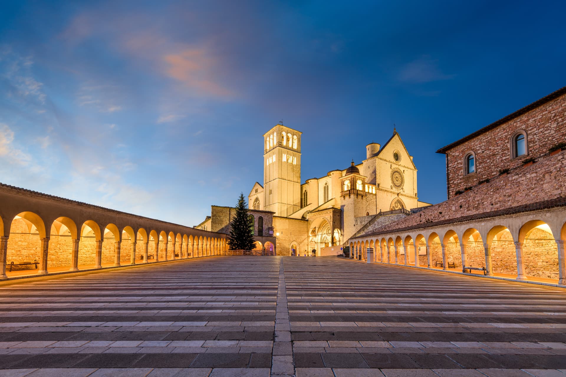 The floodlit Basilica of Saint Francis and its arcaded lower square in Assisi at dusk