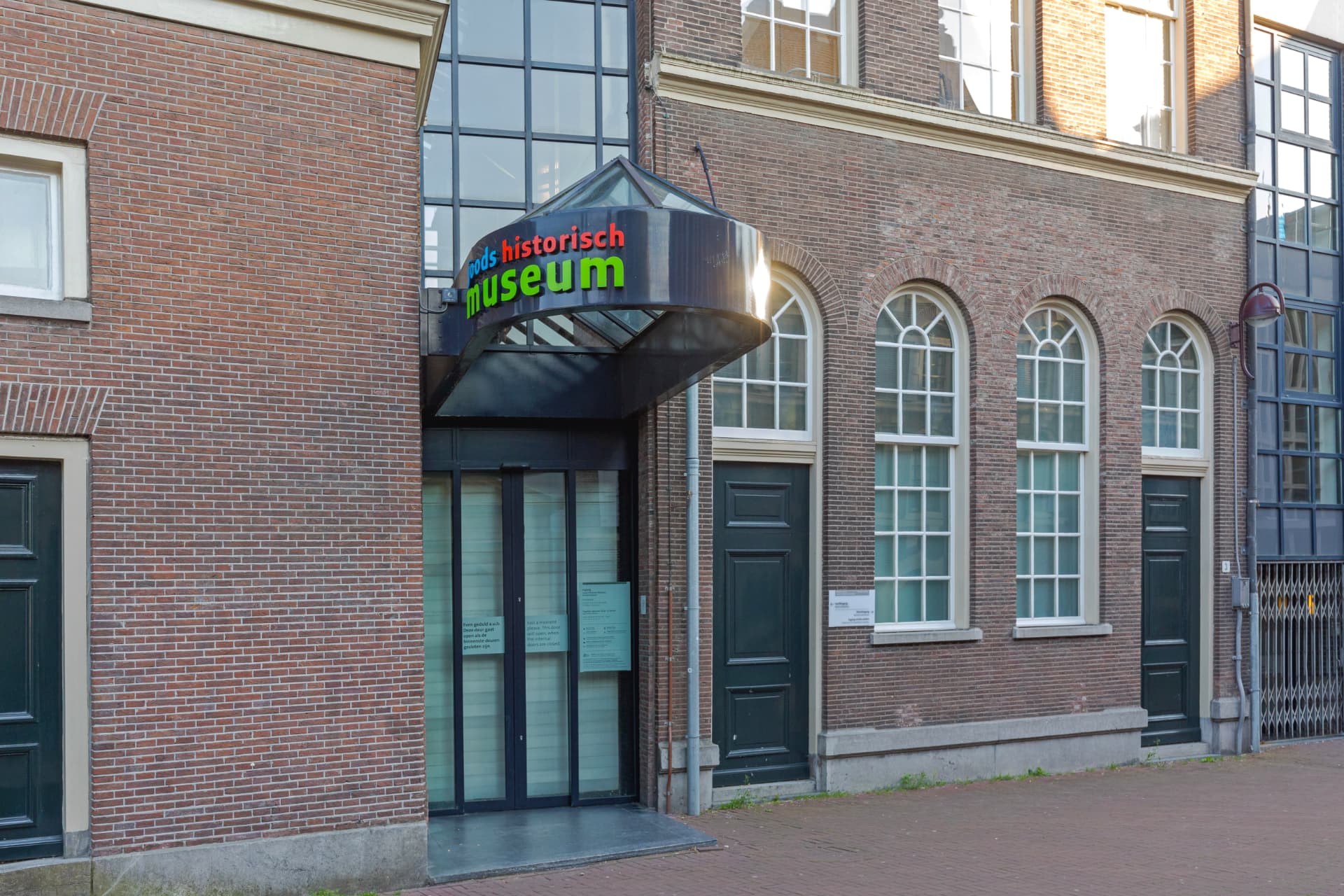 The entrance of the Jewish Museum in Amsterdam with its colourful Joods Historisch Museum sign above a glass canopy, beside arched synagogue windows
