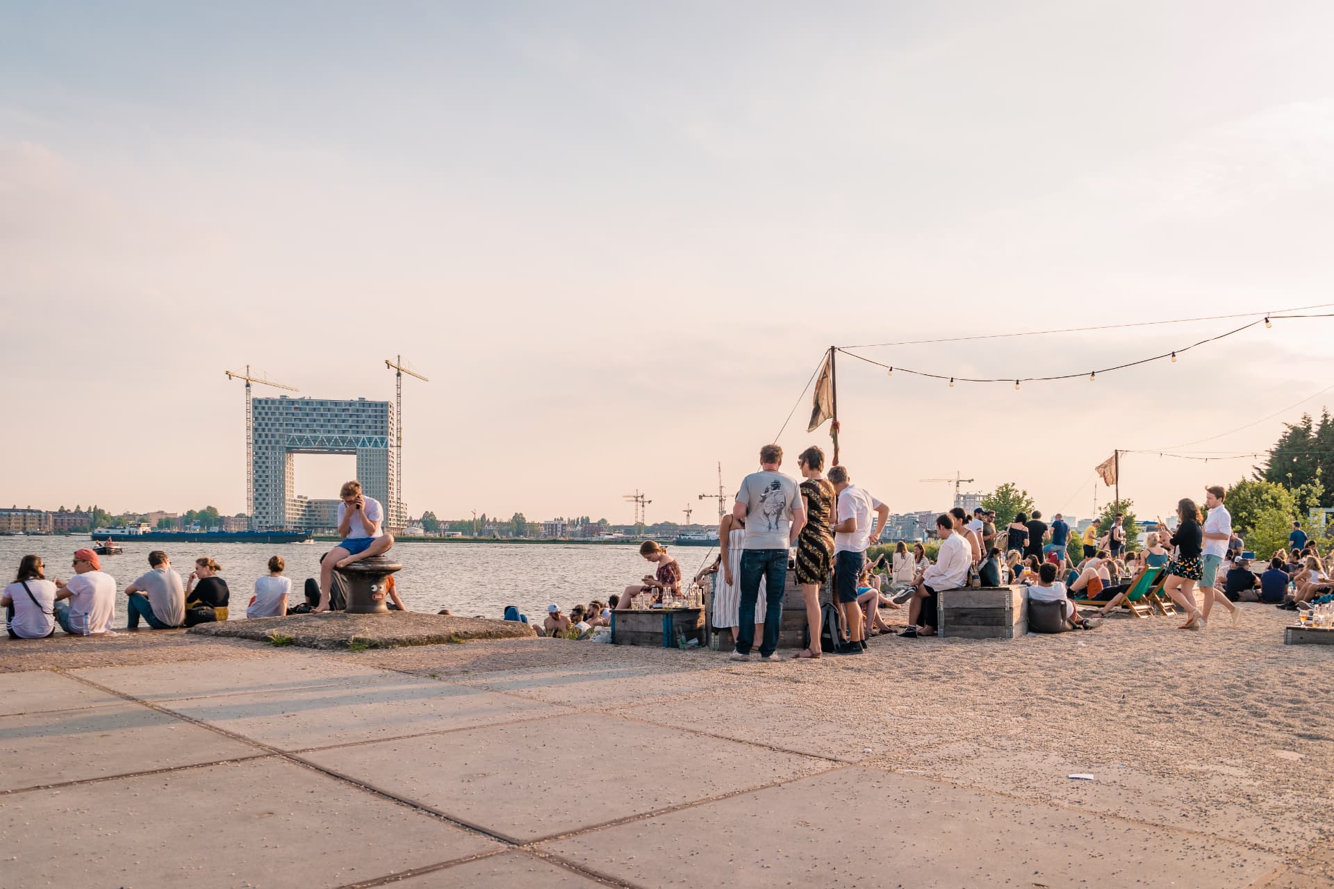 People relaxing on the NDSM Wharf waterfront in Amsterdam at sunset with the Pontsteiger building across the IJ