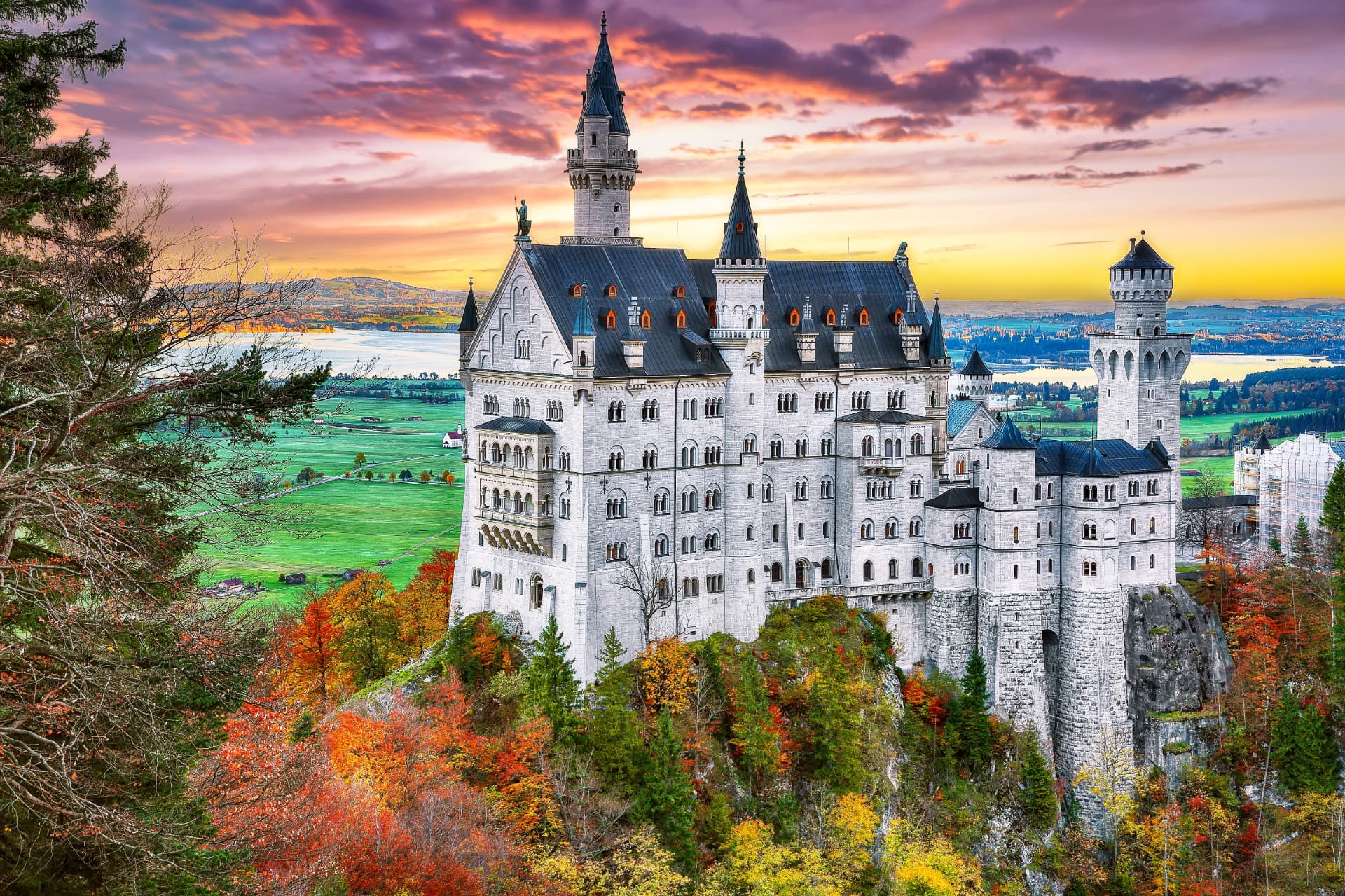Neuschwanstein Castle at sunset in autumn, surrounded by red and gold forest with the Bavarian lakes in the distance