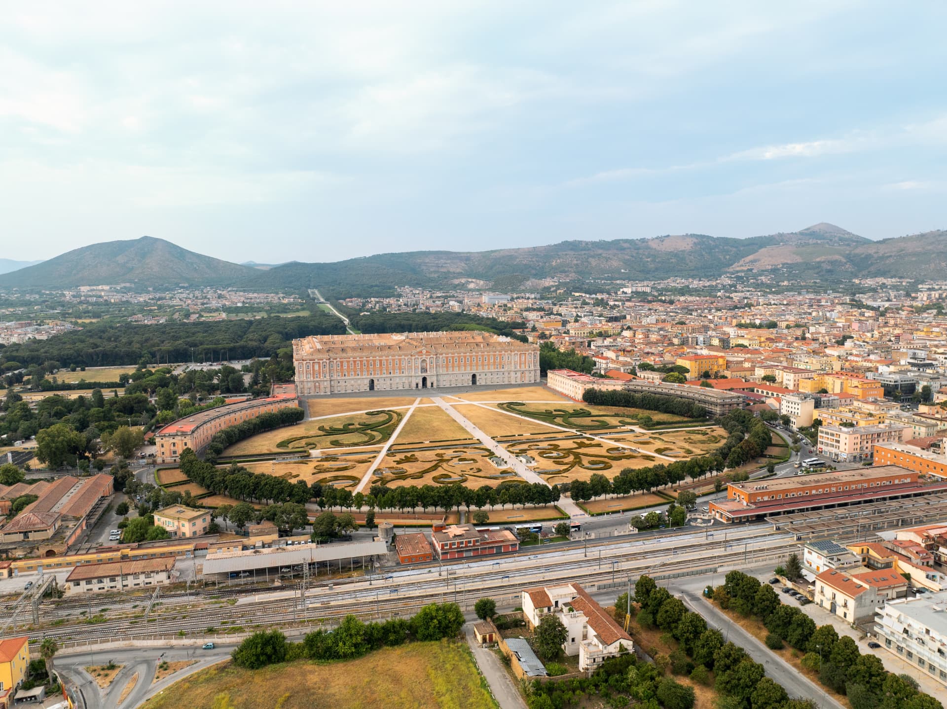 Aerial view of the Royal Palace of Caserta with the curved square in front, Caserta railway station in the foreground and the park stretching towards the hills