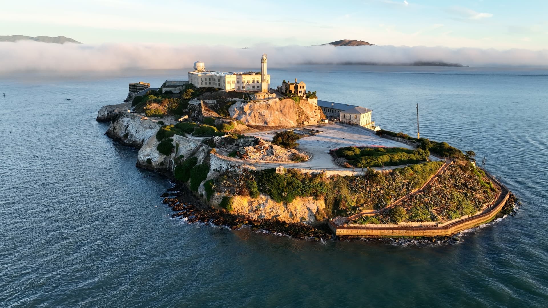Aerial view of Alcatraz Island in San Francisco Bay at sunrise, with the cellhouse and lighthouse on the ridge and fog behind