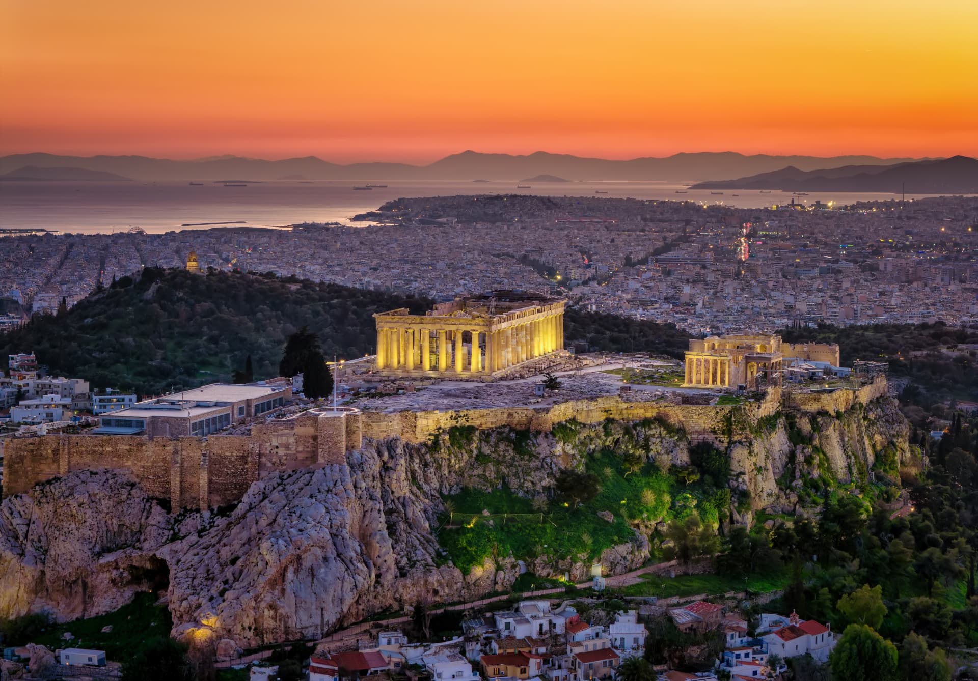 The floodlit Parthenon on the Acropolis rock above Athens at sunset, with the sea beyond
