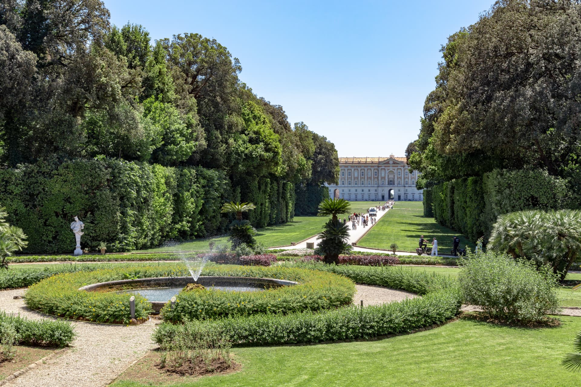 Round fountain in a garden with palm trees, framed by tall hedges, with the long avenue leading back to the palace in the distance