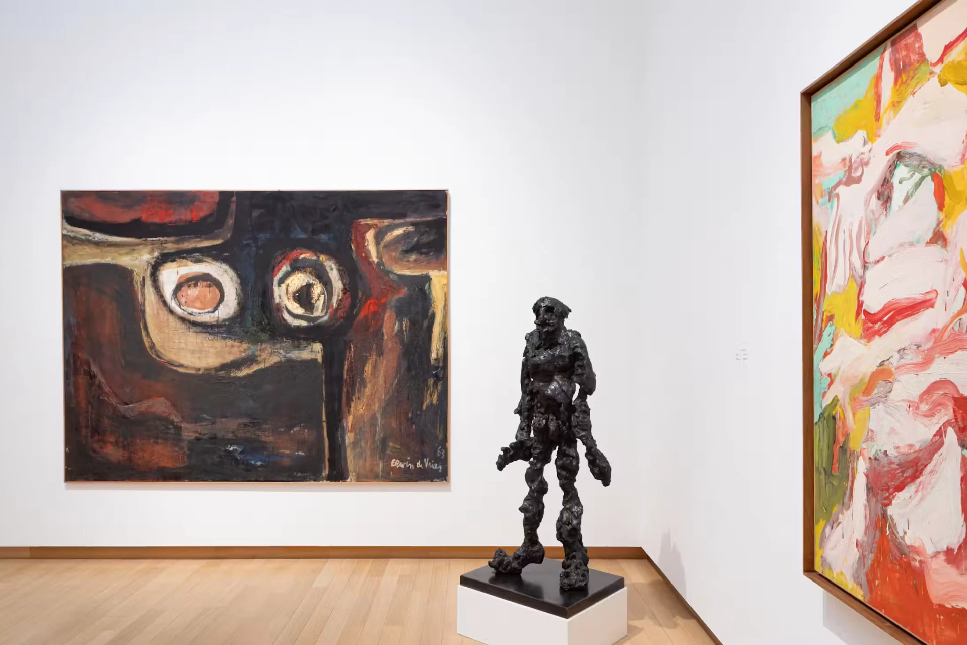 A dark bronze walking figure sculpture in a bright white Stedelijk gallery, with large abstract paintings on the walls