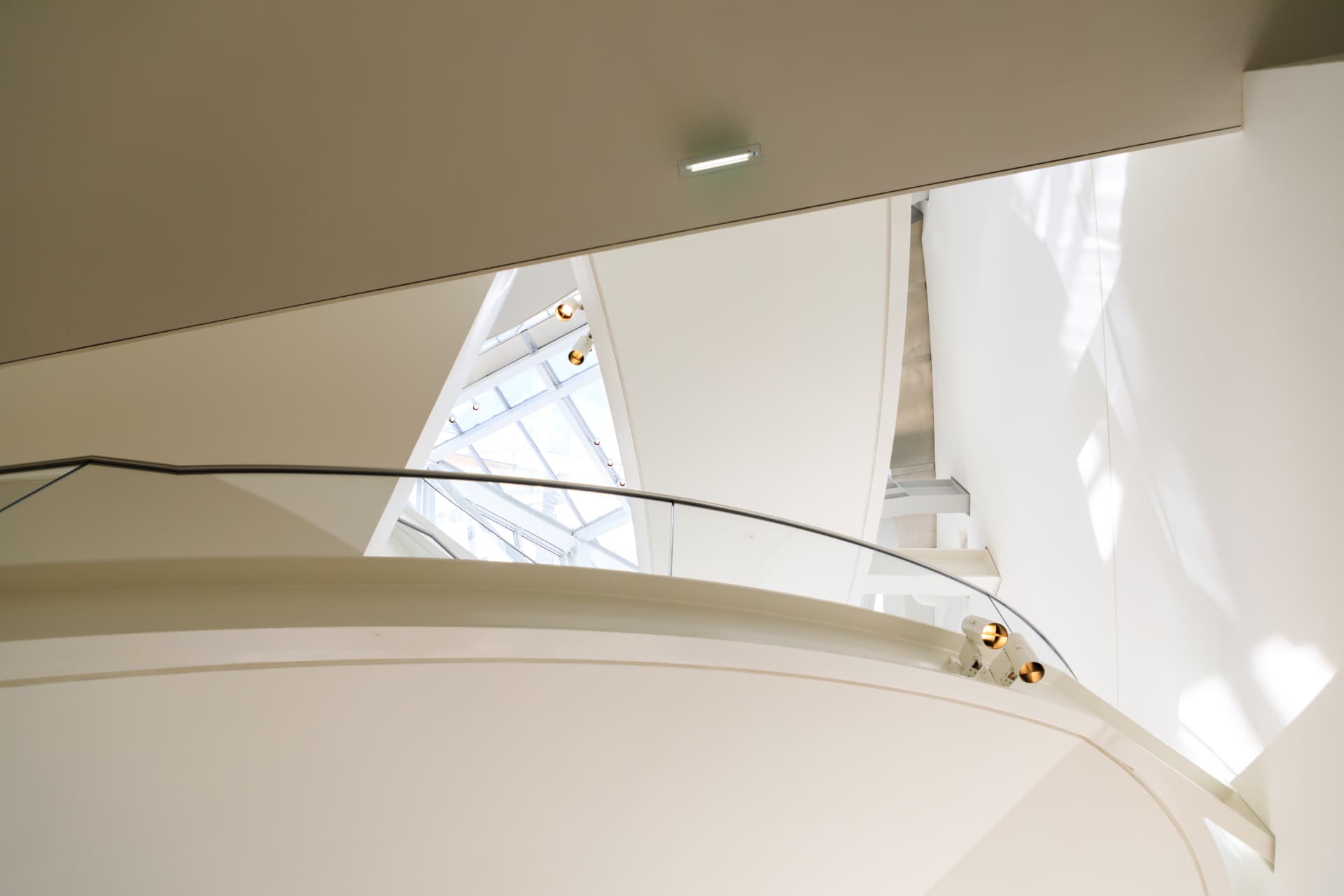 A curving white balcony and glass balustrade inside the Fondation Louis Vuitton, with daylight coming through a triangular gap between the glass sails