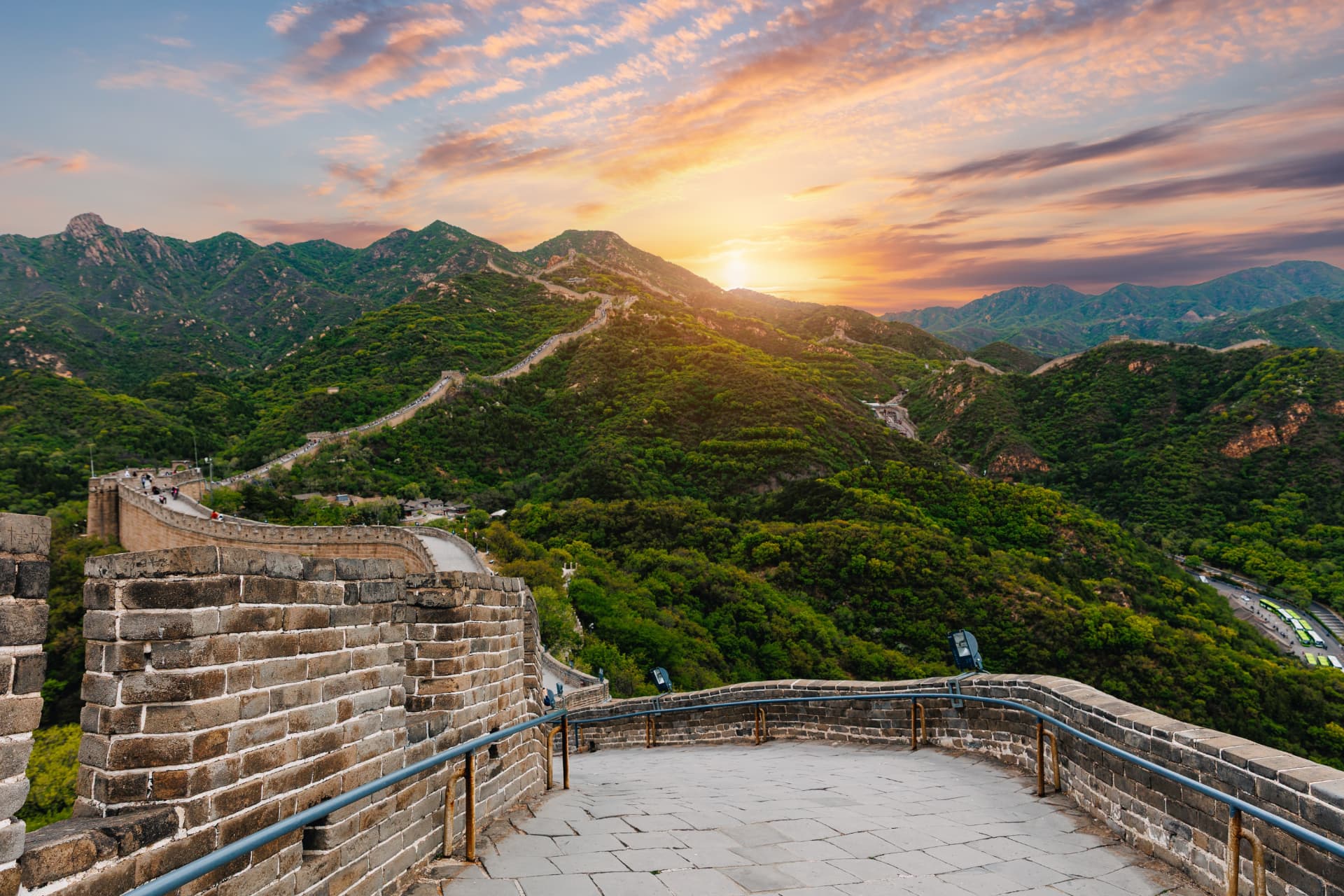 A stone viewing platform on the Great Wall at sunset, with the wall running away over green ridges into the distance