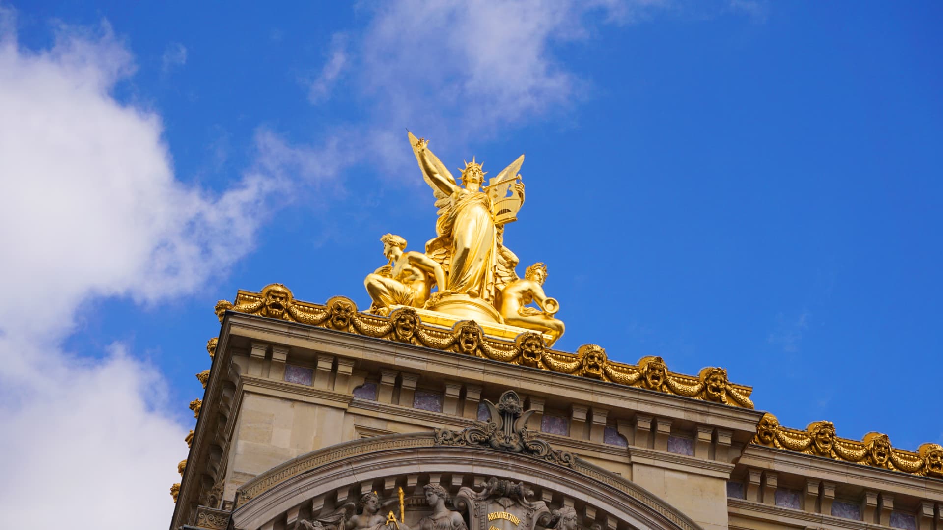 Gilded statue group on the roofline of the Palais Garnier against a blue sky