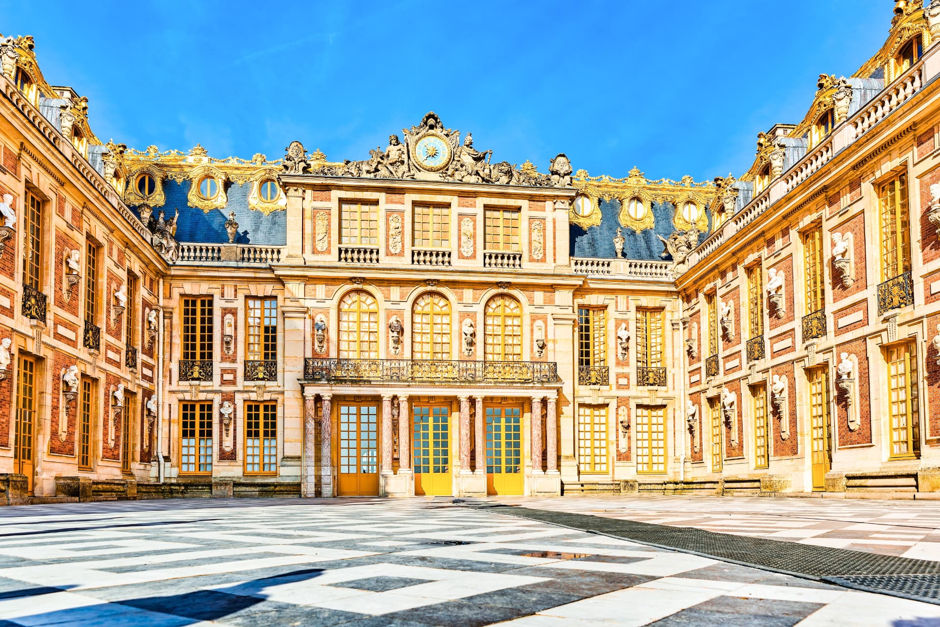 The marble courtyard and richly decorated facade of the Palace of Versailles under a clear blue sky