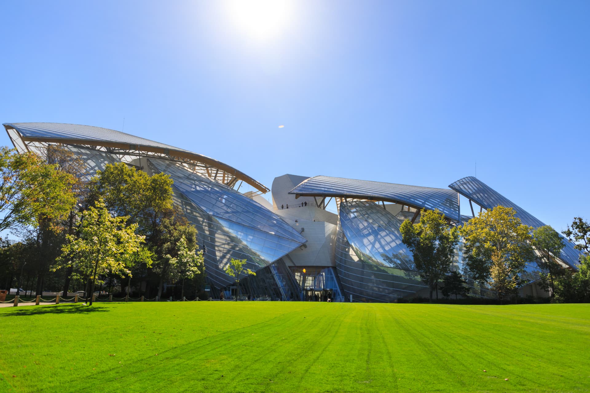 The Fondation Louis Vuitton seen across a green lawn, its glass sails catching the sun above the trees of the Bois de Boulogne