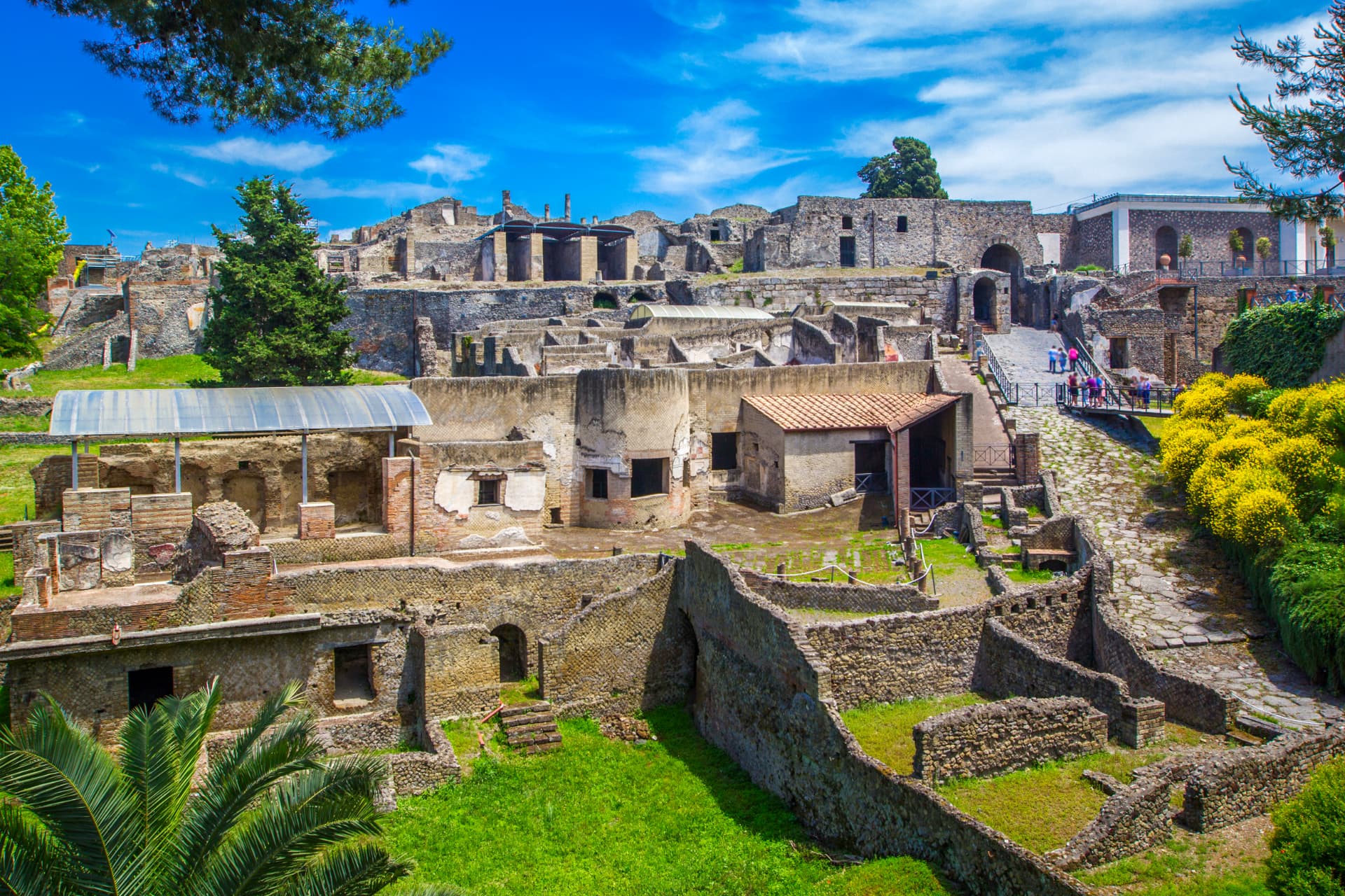 Excavated streets and ruined walls of Pompeii near the main entrance, with visitors entering the archaeological park