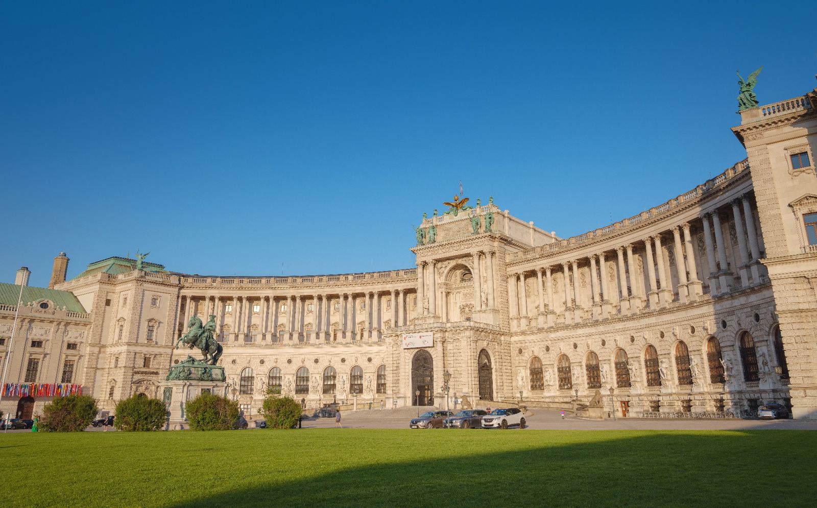 Hofburg Palace in Vienna seen from Heldenplatz with its curved Baroque facade, tall columns, bronze equestrian statue and green winged statues under a clear blue sky